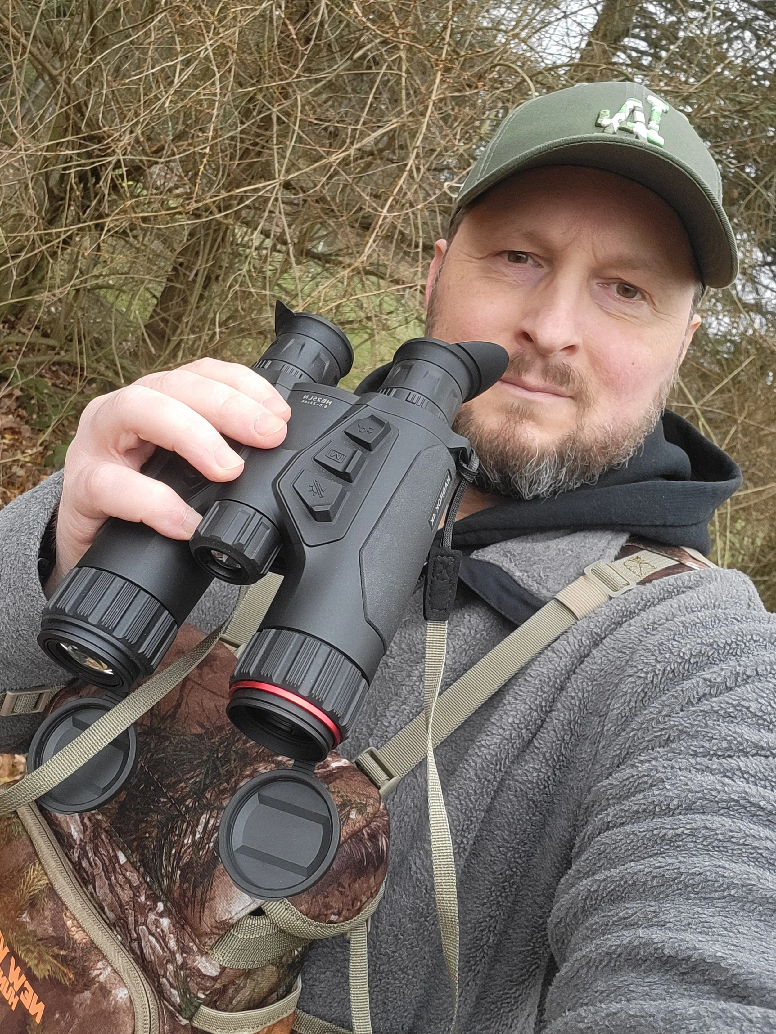 Ein Fotograf mit Cappy und Jacke, hält ein multispektrales Fernglas und steht im Wald.
