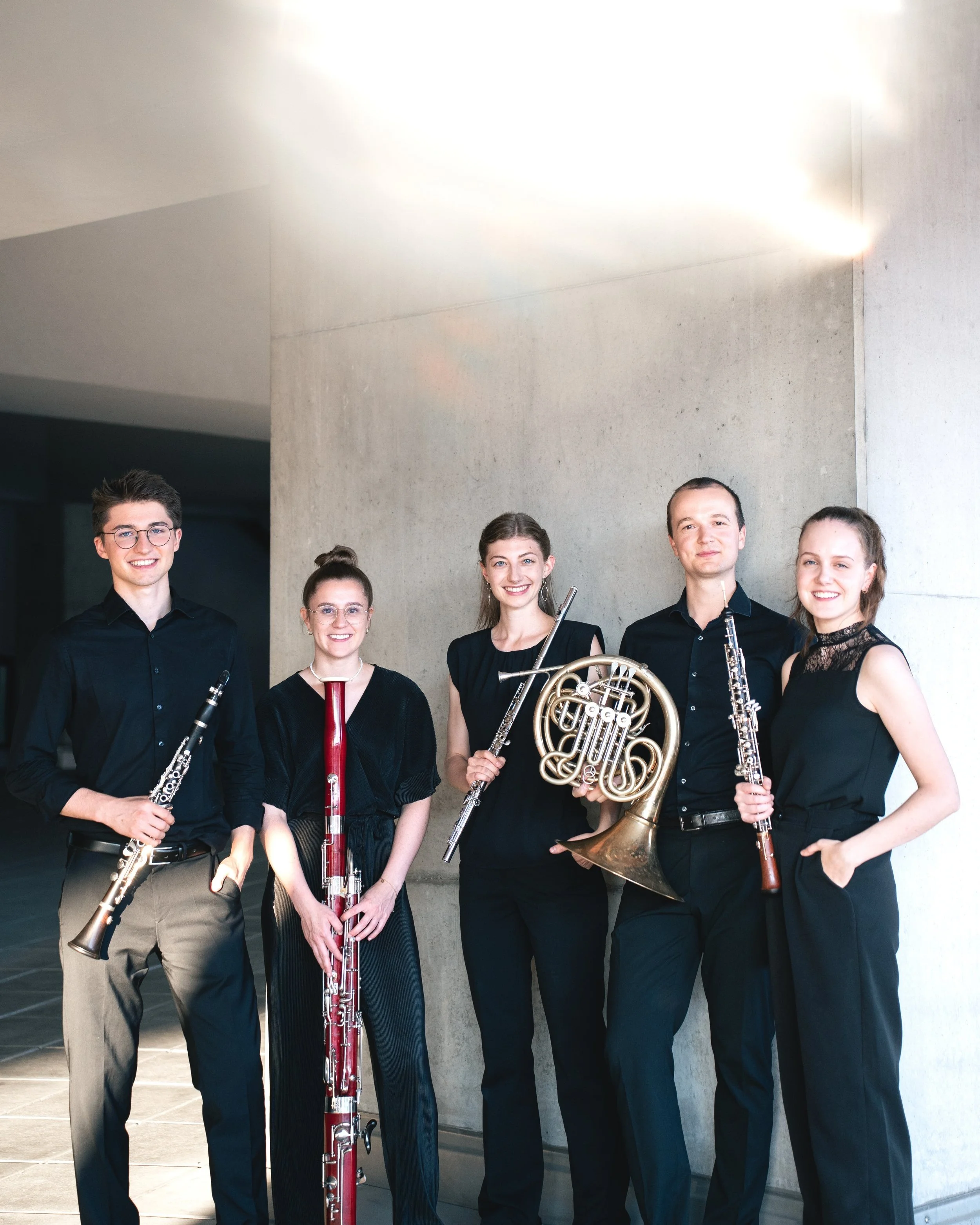 Fünf junge Musiker stehen mit ihren Blasinstrumenten vor einer Betonwand. Es sind drei Frauen und zwei Männer, alle tragen schwarze Kleidung. Sie sind lachend und bereit für eine Aufführung.