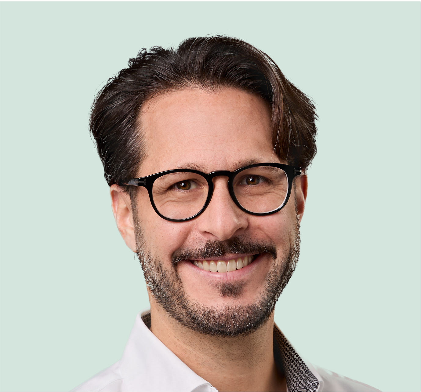 Porträt eines lächelnden Mannes mit dunklen Haaren, Brille und einem weißen Hemd gegen einen hellgrünen Hintergrund.