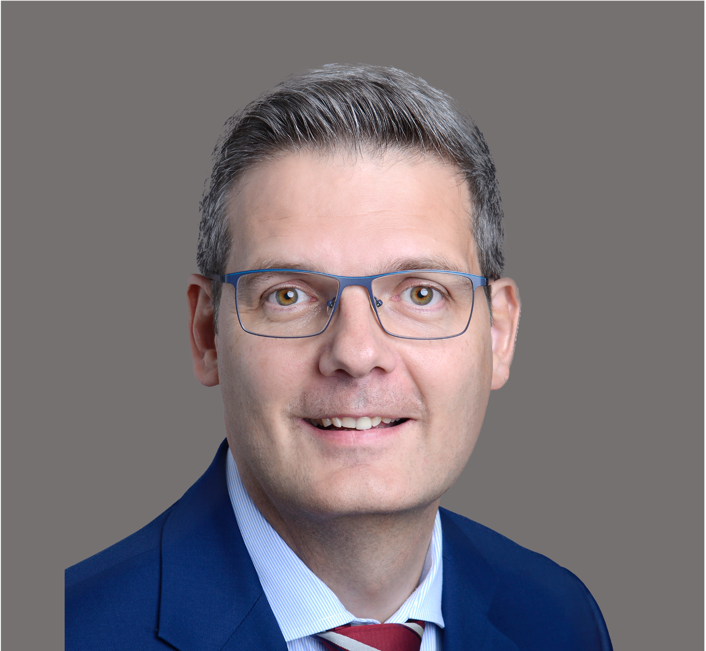 Porträt eines Mannes mit dunklem Haar, Brille, Anzug und Krawatte vor beige Hintergrund.