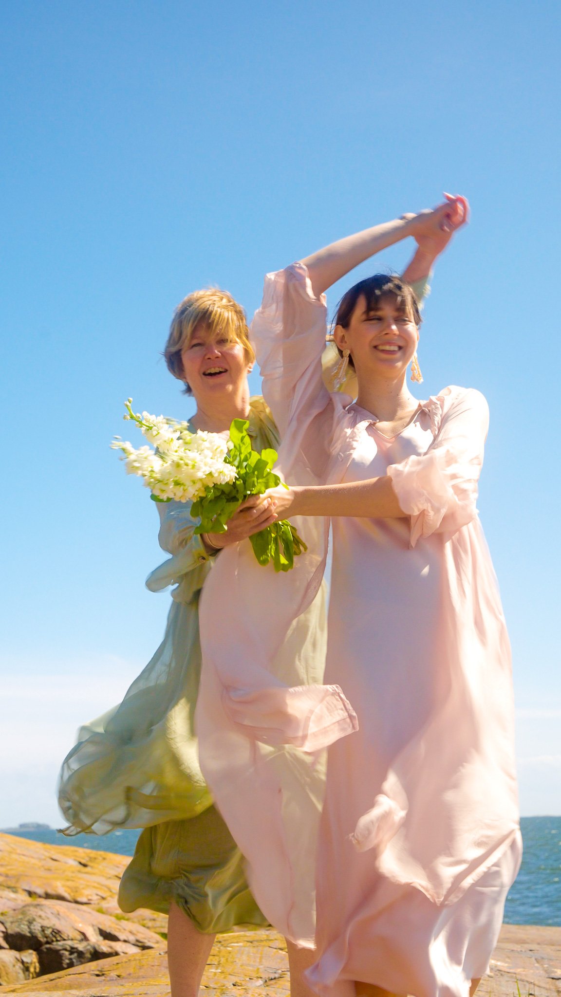 Two women, one holding a bouquet of white flowers, are running outdoors on a sunny day near water, smiling and enjoying themselves.