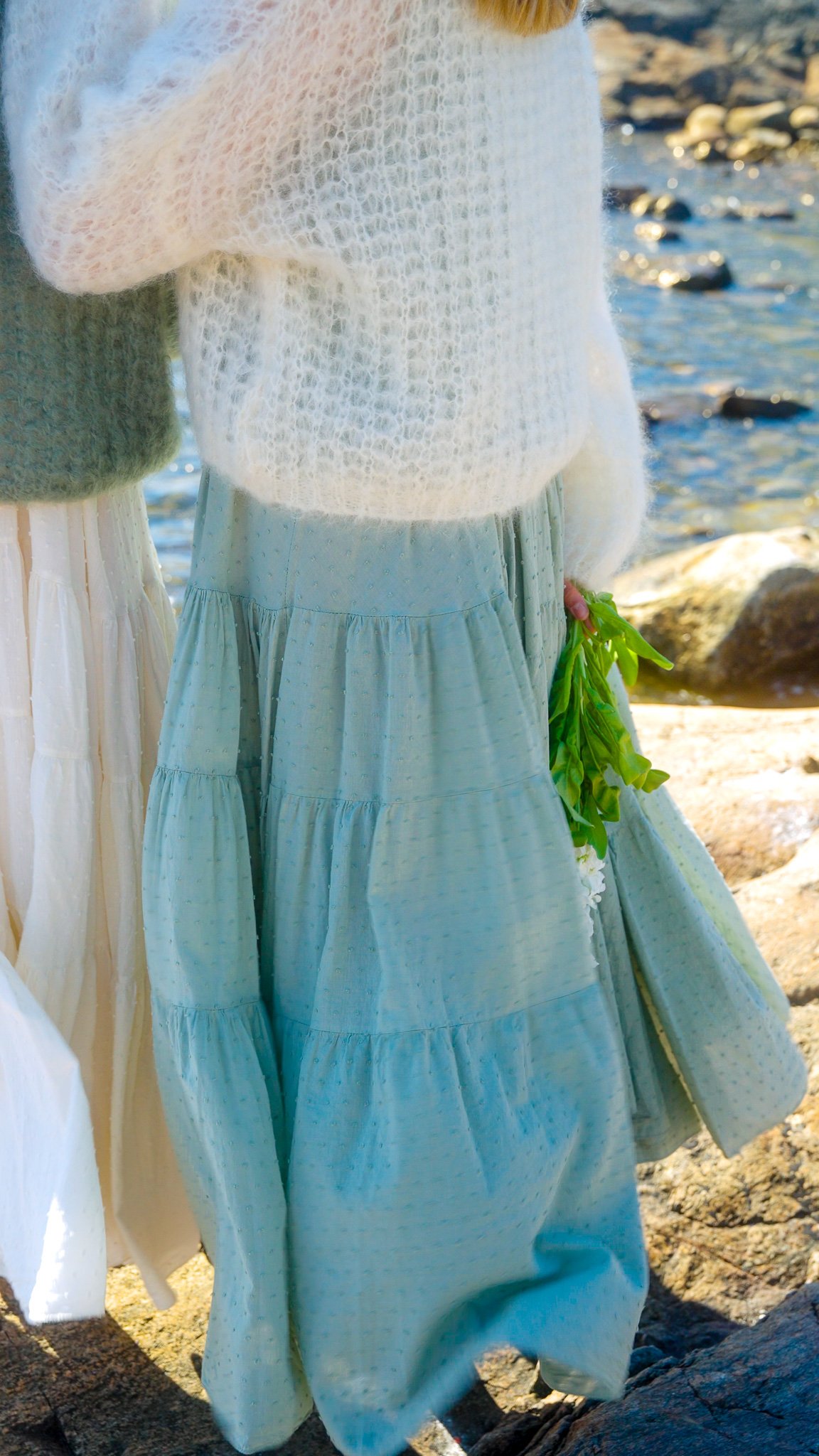 Close-up of two women standing by rocks and water, one wearing a white or cream skirt and a fuzzy sweater, holding green plants, near a rocky shoreline.