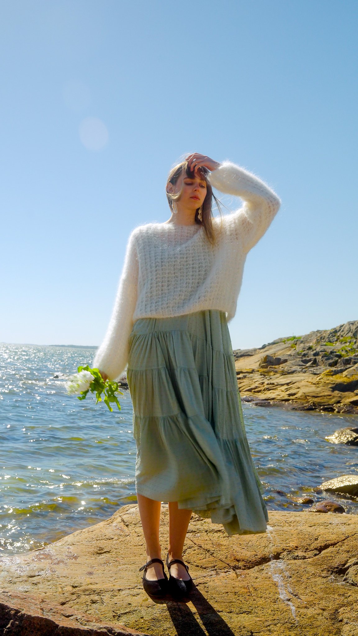 Woman standing on rocks by the water under clear blue sky, wearing a white sweater and gray skirt, holding white flowers.