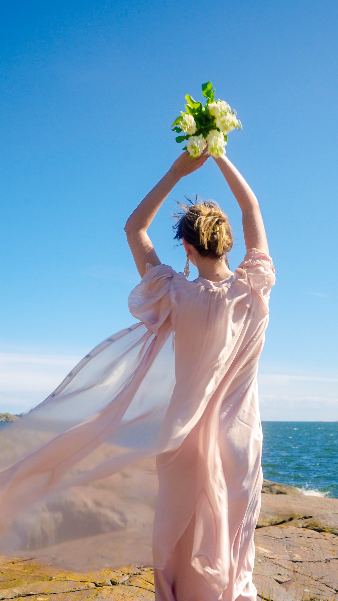 A woman dressed in a flowing, light pink dress standing on a rocky shoreline with the ocean and a blue sky in the background, holding a bouquet of white flowers above her head.
