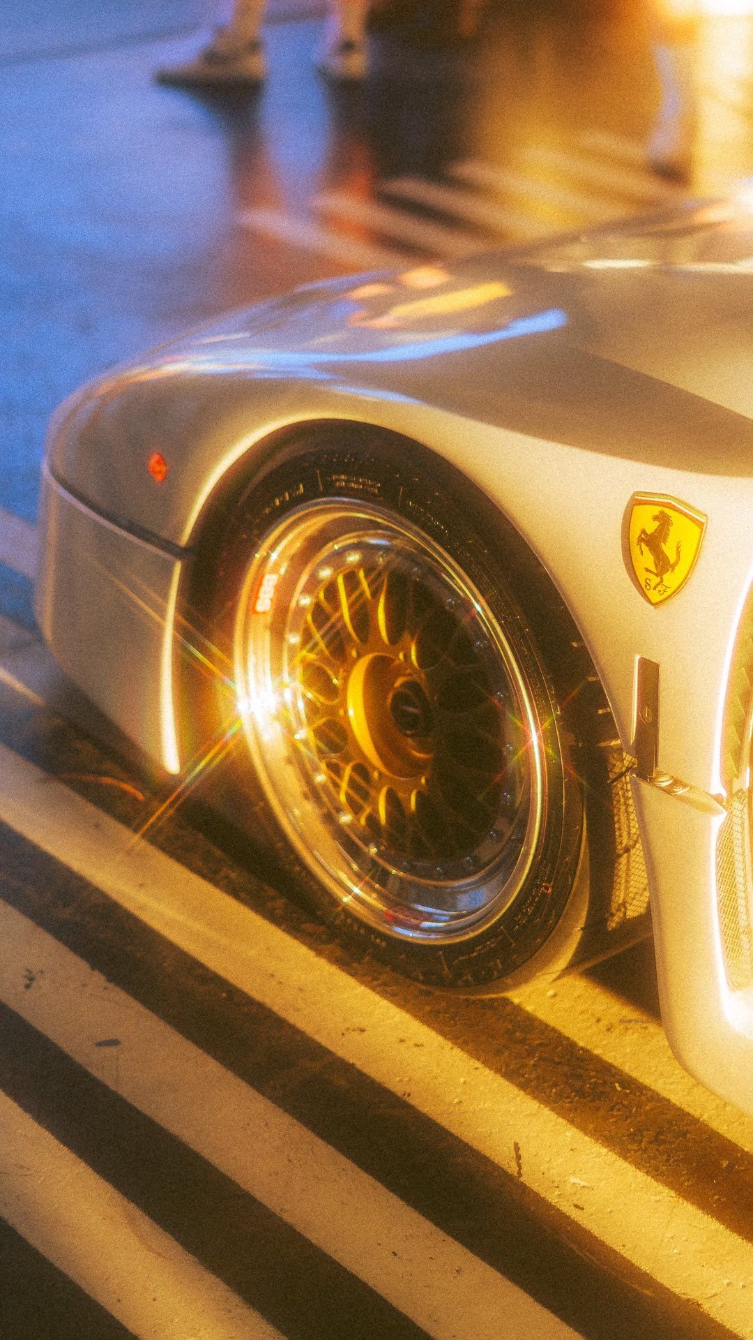 Close-up of a silver Ferrari sports car with a yellow ferrari logo on the front, parked on street at night with reflections and glare from lights.