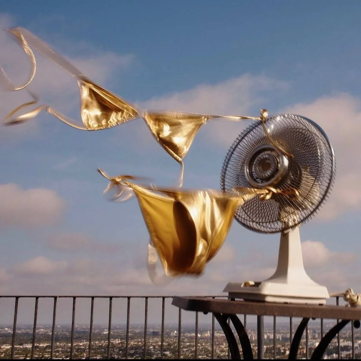 Golden bikini top and bottom hanging on a clothesline next to a large electric fan on a balcony with a city view.