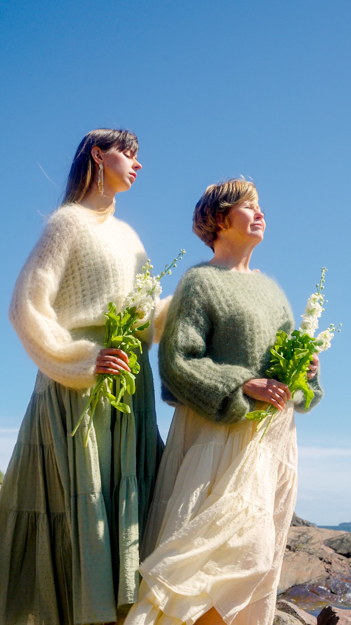 Two women standing outdoors by rocks holding white flowers, wearing sweaters and long skirts, with a bright blue sky in the background.