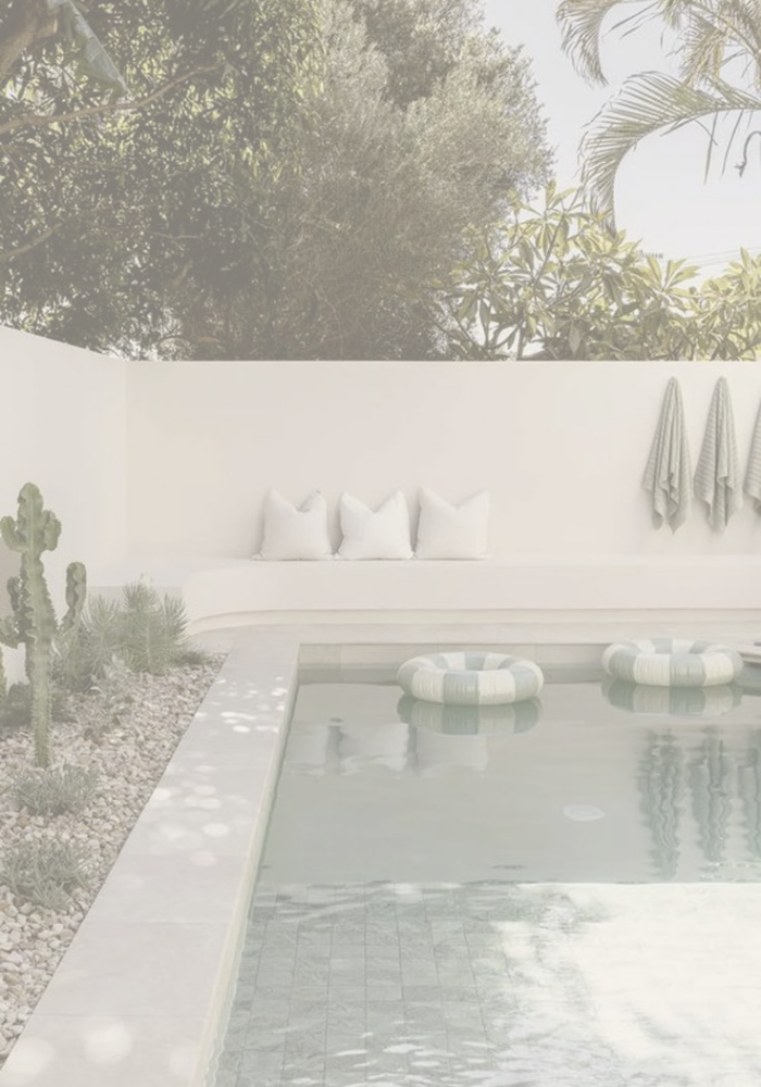 A serene backyard with a swimming pool, pool floats, a white bench with three pillows, wall-mounted towels, and desert plants including cacti, with a mural of trees and foliage in the background.