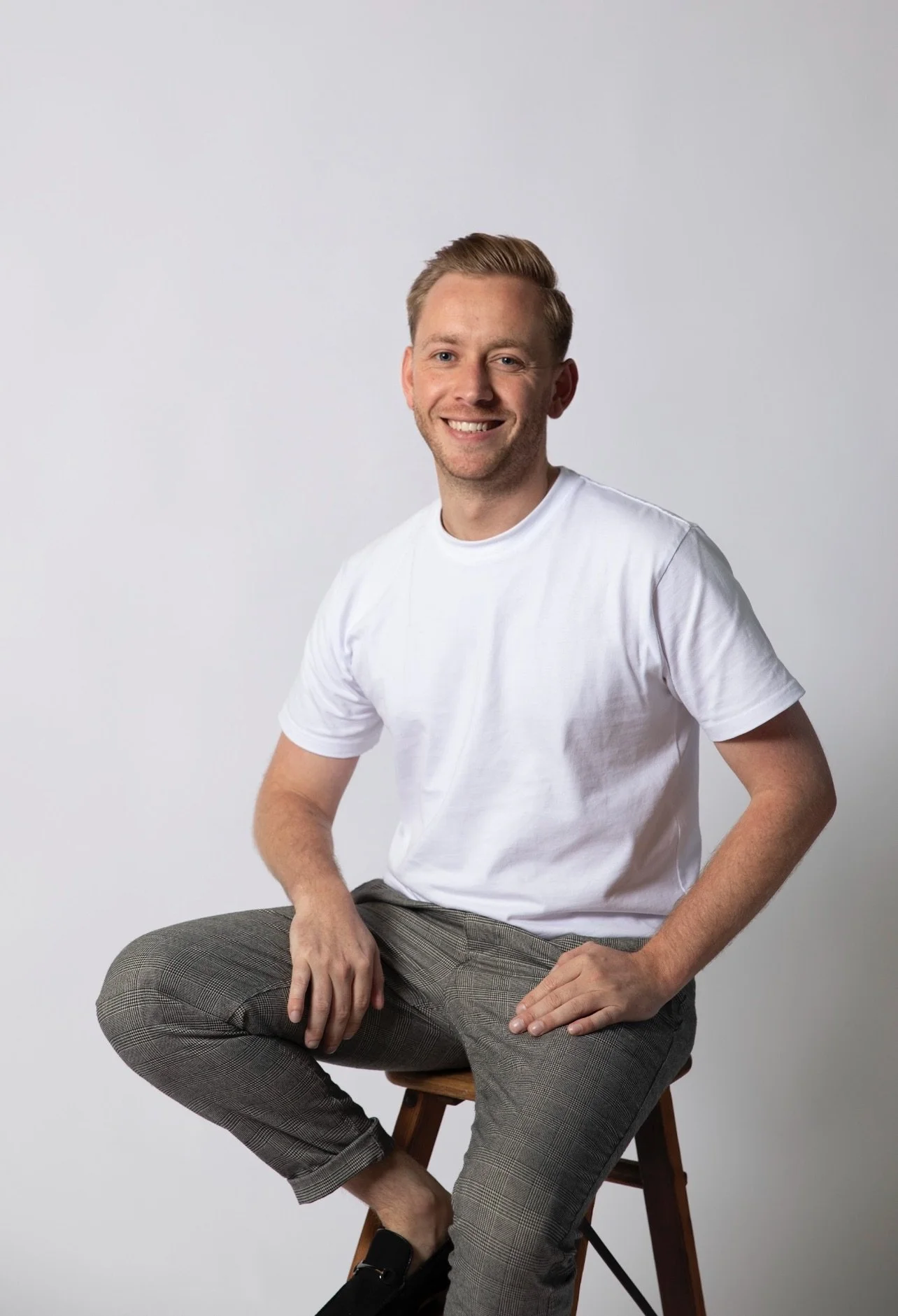 A smiling man with short blond hair, wearing a white t-shirt and plaid pants, sitting on a stool against a plain white background.