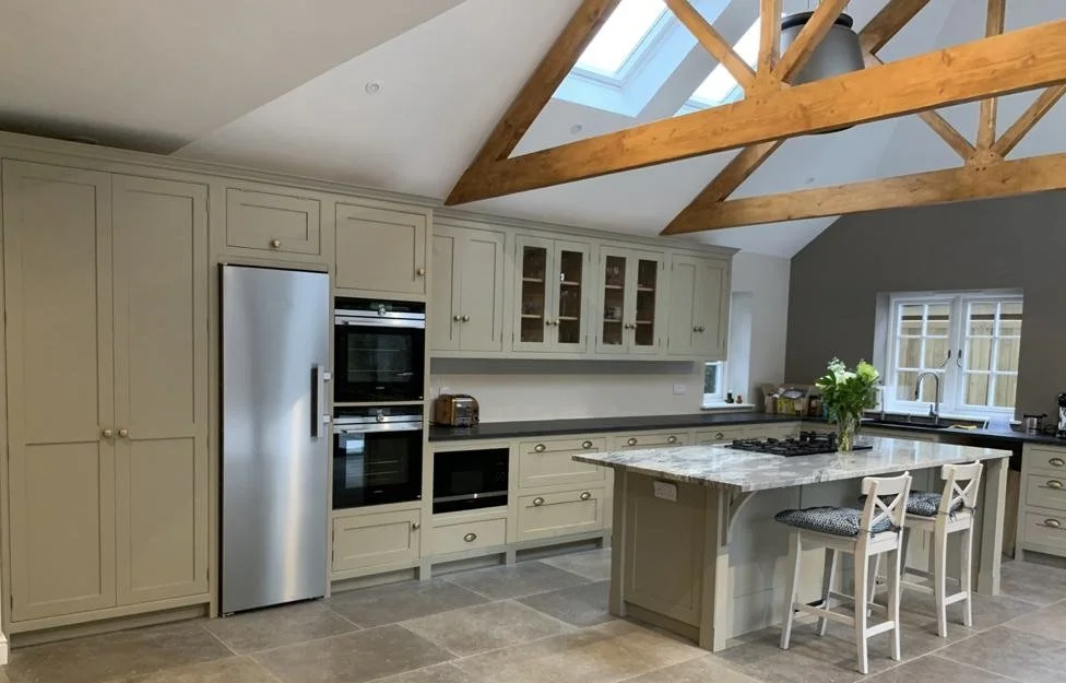 Modern kitchen with cream-colored cabinets, a stainless steel refrigerator and oven, a marble island with two chairs, a skylight, and wooden beams on the ceiling.