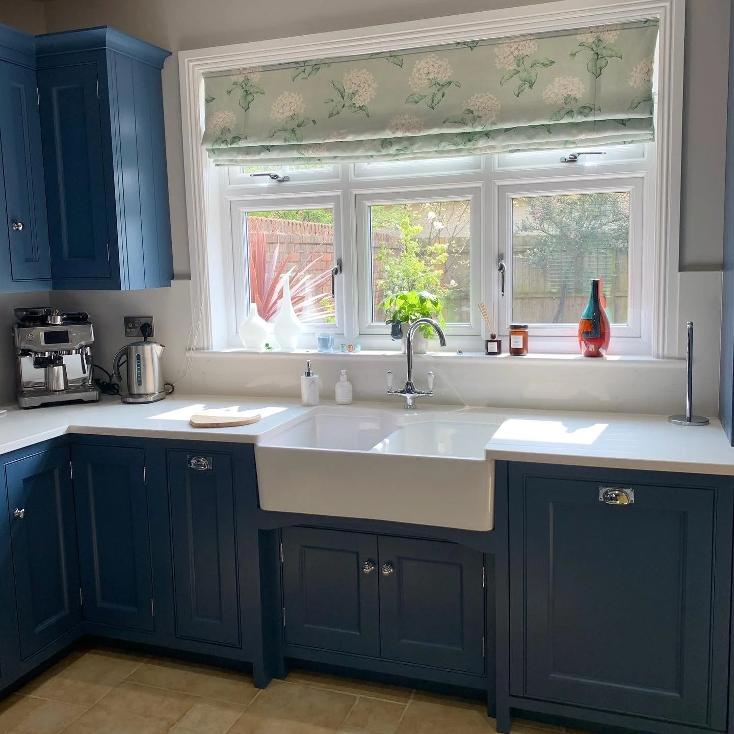 Kitchen with blue cabinets, white countertop, farmhouse sink, and window above it. The window has a floral roman shade, with decorative vases and plants on the windowsill. A coffee machine and electric kettle are on the left side of the counter.