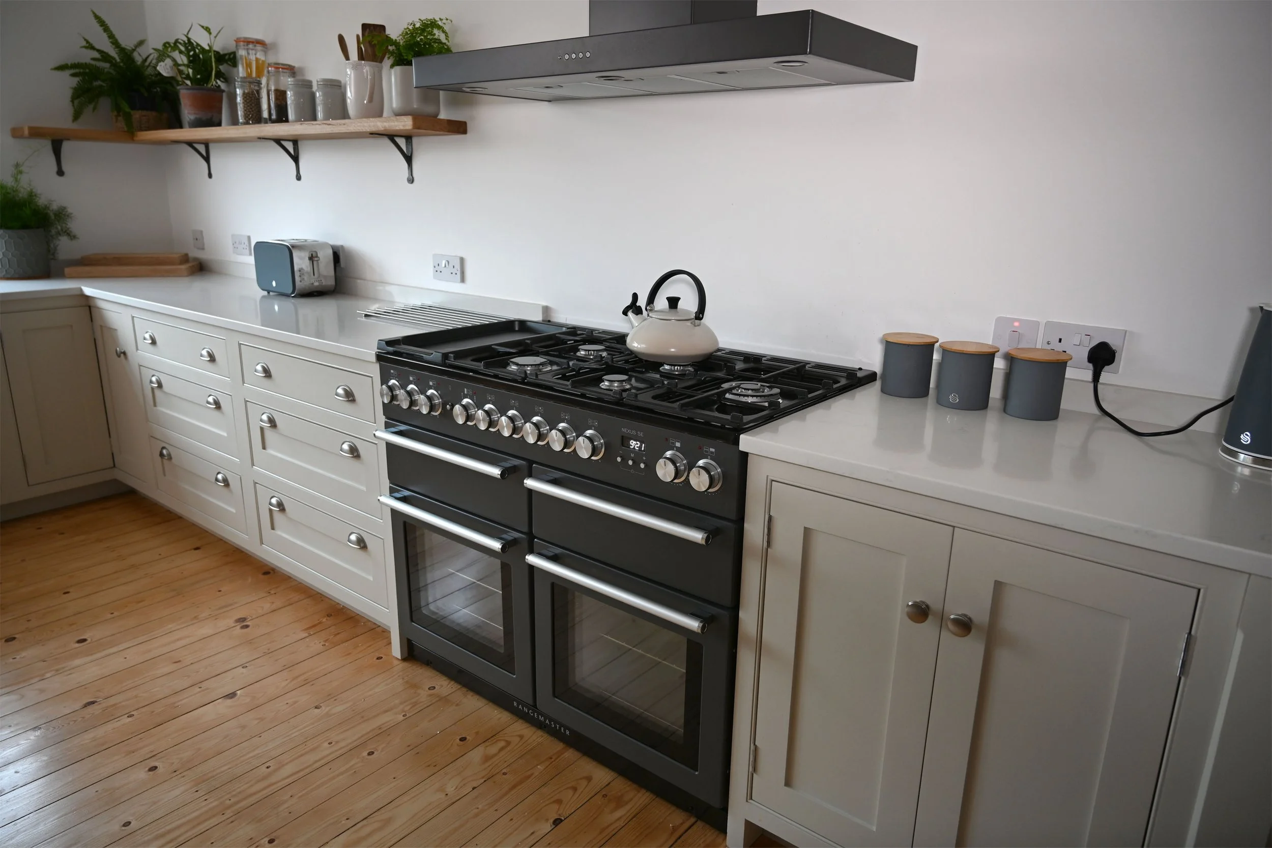 Kitchen with cream-colored cabinets, a black and stainless steel oven and stove, a white kettle on the stove, open wooden shelves with plants and jars, countertop with toaster, and four gray canisters with wooden lids, wooden flooring, and a white wa