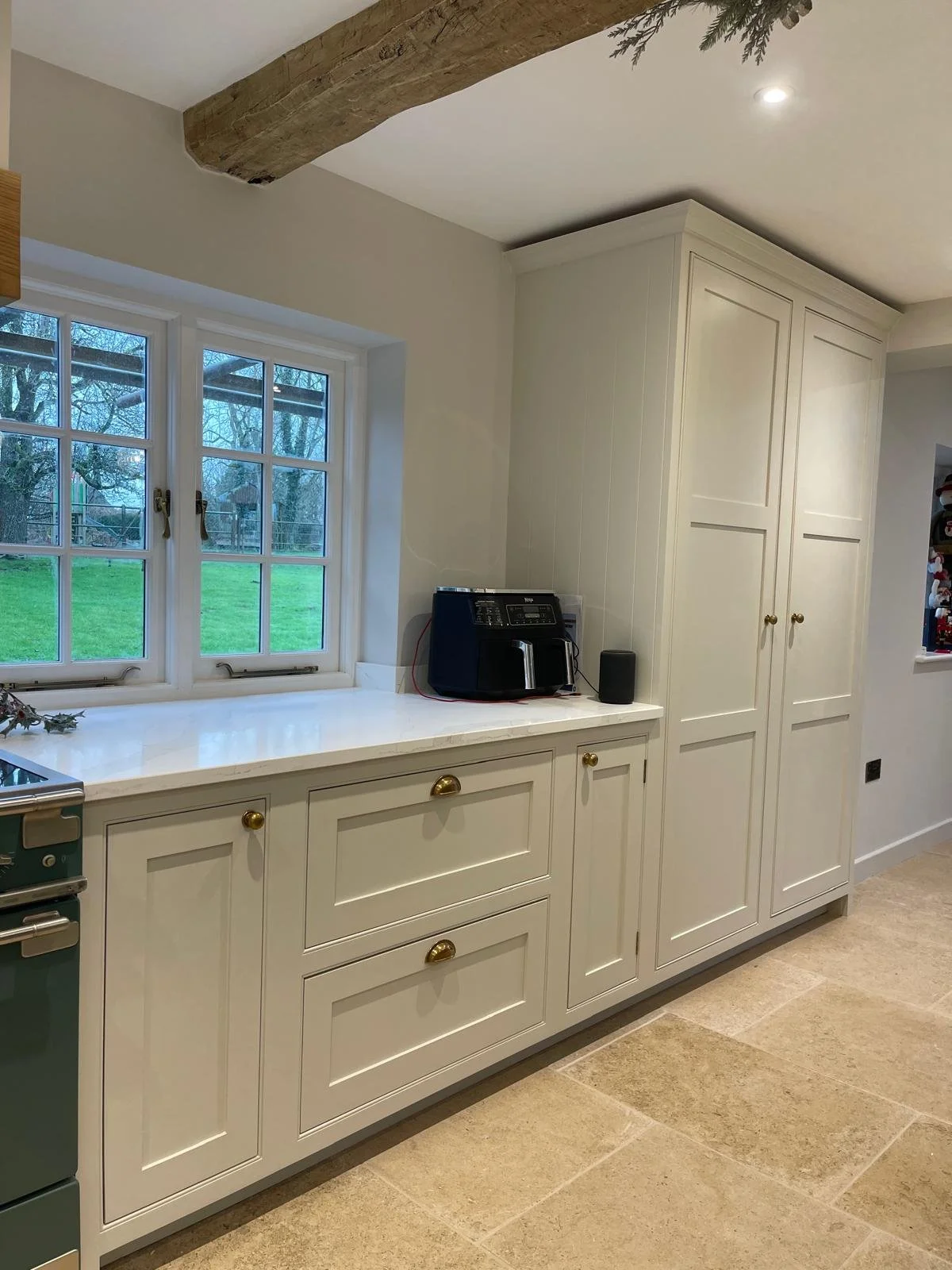 White kitchen cabinet with gold knobs beneath a window, with a black coffee maker and speakers on the countertop, crafted from wood with a rustic beam on the ceiling, overlooking a green backyard.