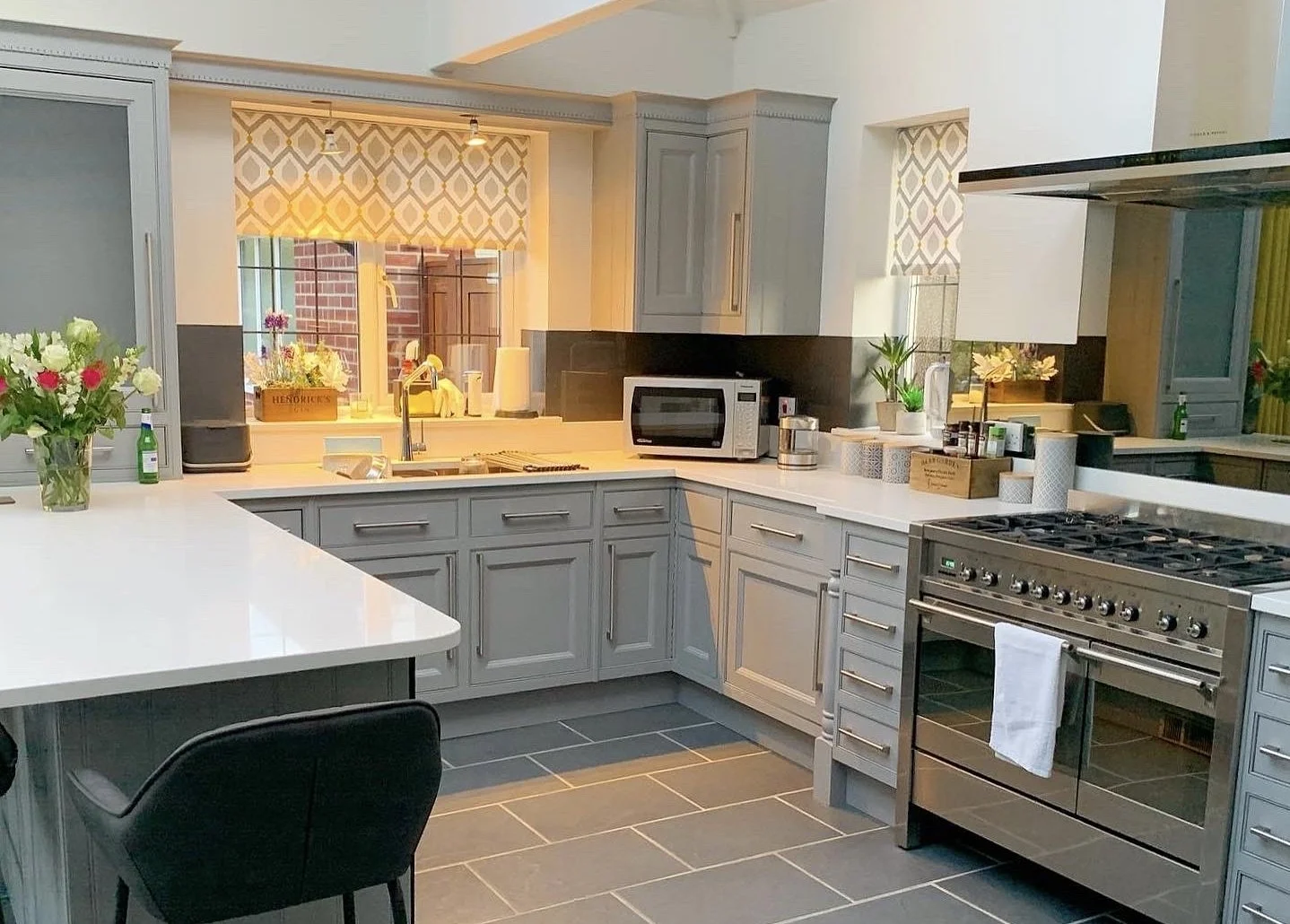Modern kitchen with gray cabinets, stainless steel appliances including a stove and microwave, a white countertop, flowers in a glass vase, and a window with patterned shades.