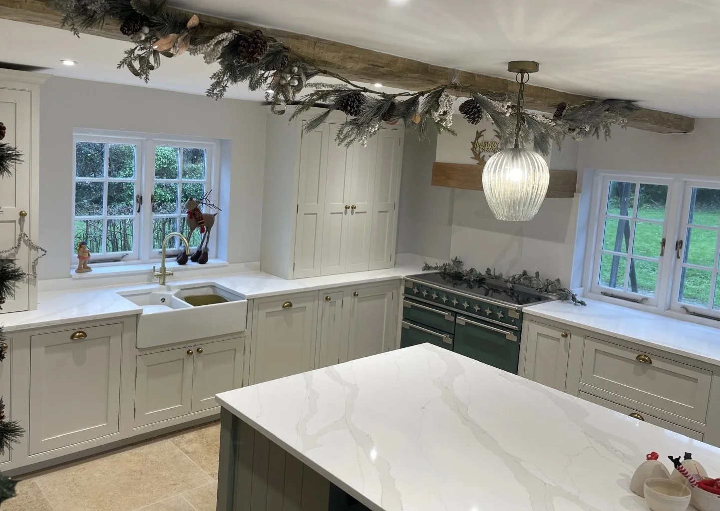 Kitchen decorated for Christmas with white cabinets, a marble countertop, two windows, a farmhouse sink, a green stove with star decorations, and festive garland hanging from the ceiling.
