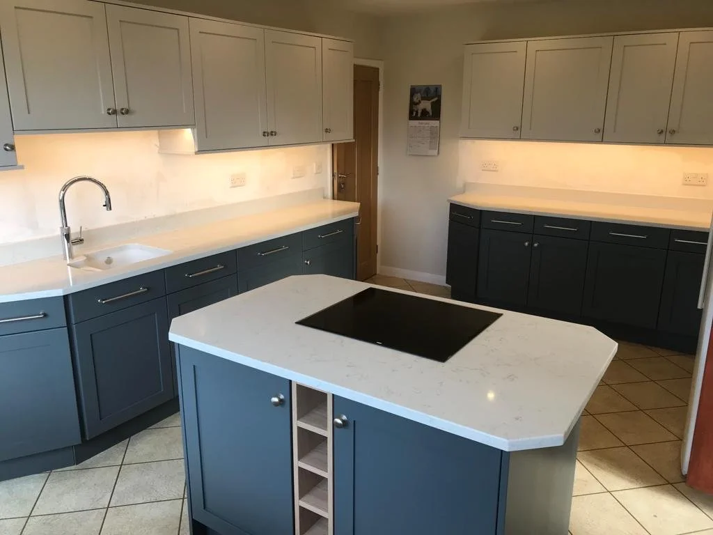 Modern kitchen with blue and white cabinetry, a white countertop, a built-in black cooktop on a central island, a white sink with a silver faucet, tiled floor, and a calendar on the wall.