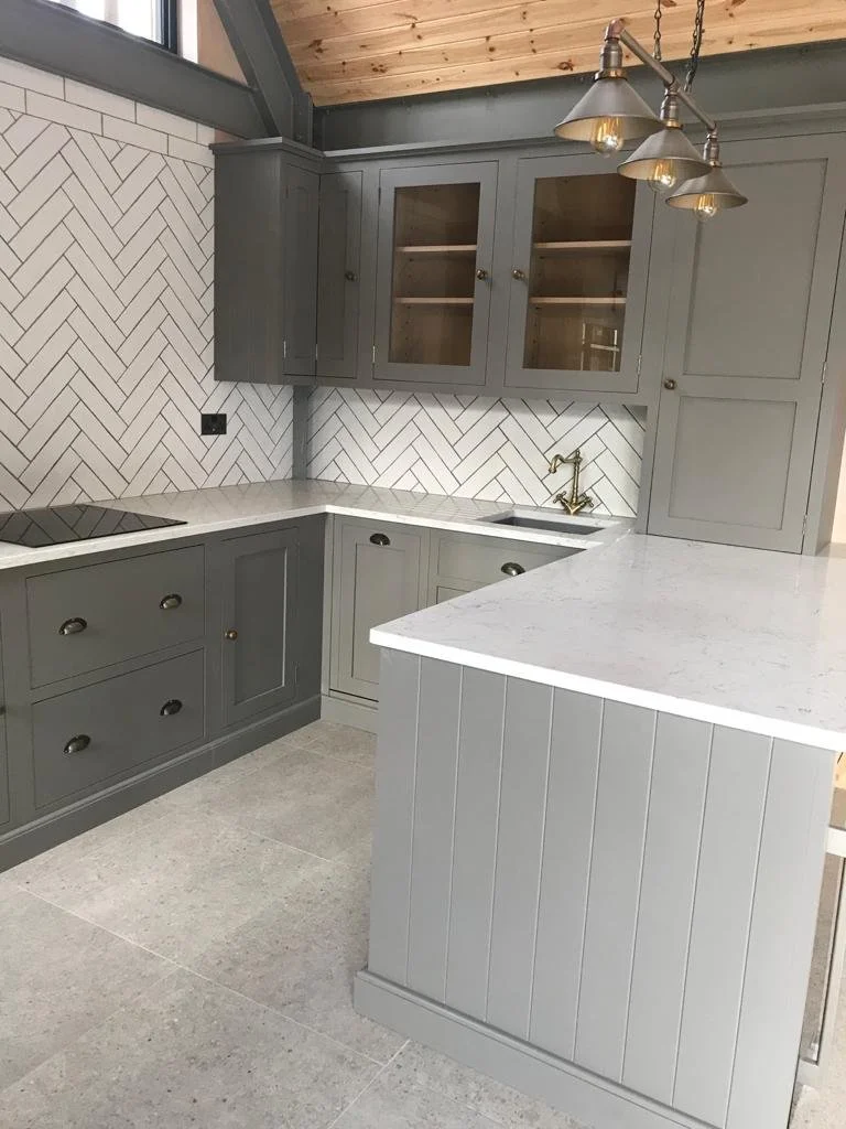 Modern kitchen with gray cabinets, white marble countertops, herringbone patterned backsplash, gold faucet, and industrial style pendant light fixtures.