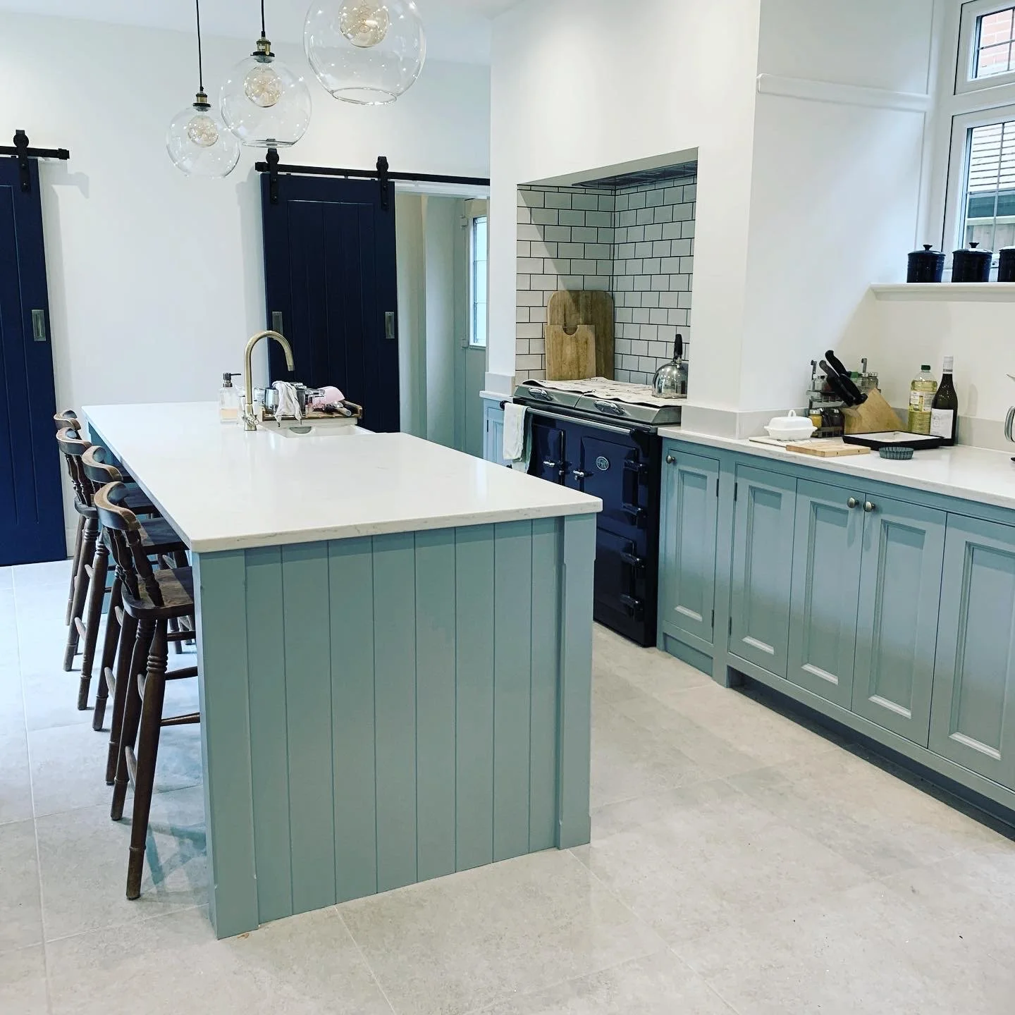 Modern kitchen with a large island with white countertop and blue trim. Blue cabinetry, a black stove, and pendant lights with glass globes hanging from the ceiling. Black sliding barn doors and windows with white frames.