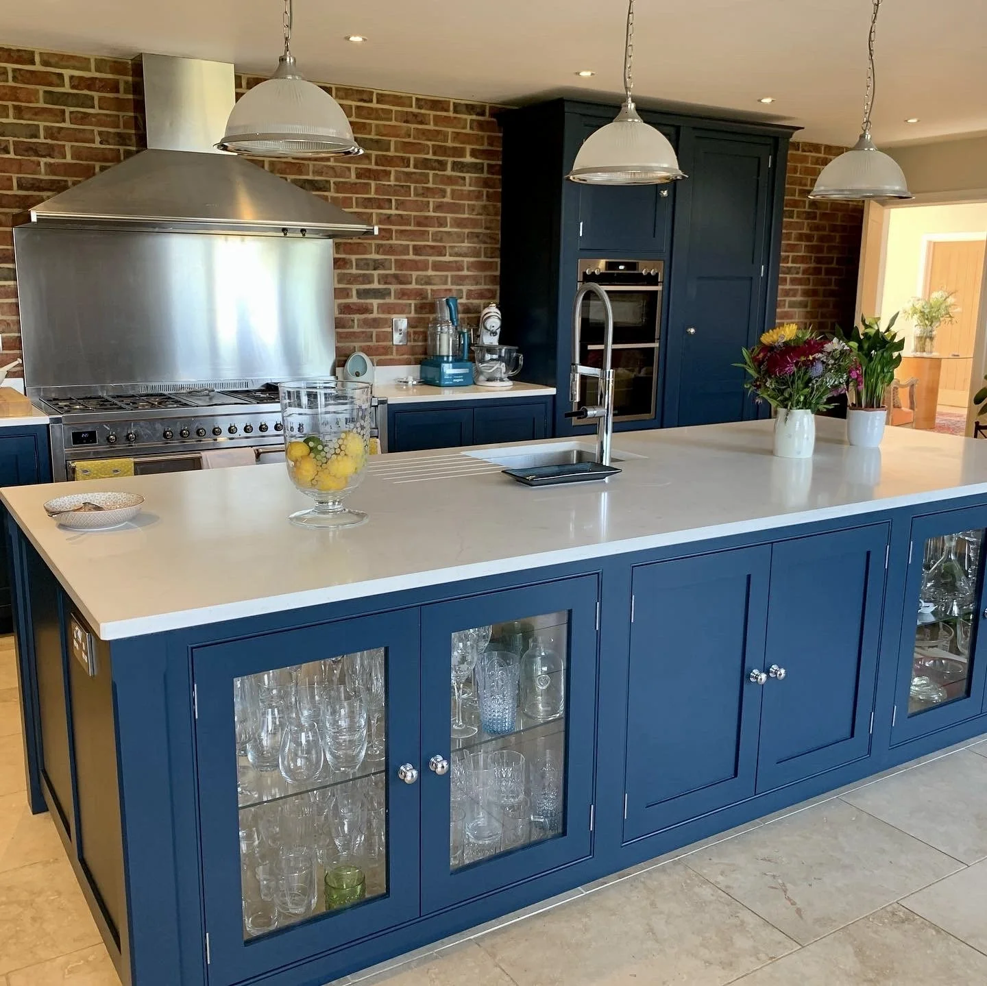 Kitchen island with blue cabinetry, white countertop, glassware inside cabinets, a sink with a tall faucet, and a vase with flowers. In the background, stainless steel stove, brick wall, navy cabinets, and kitchen appliances.