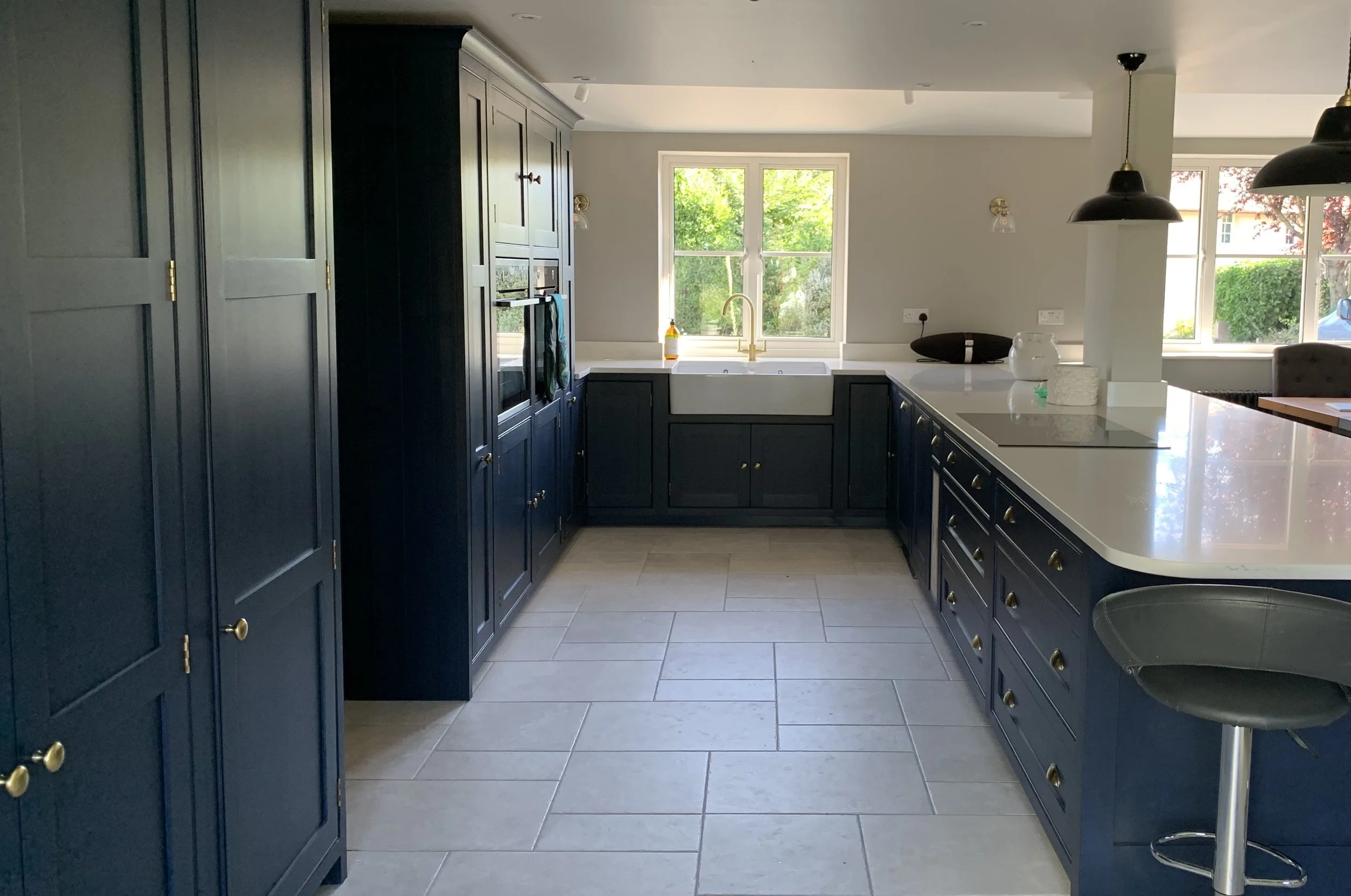 Modern kitchen with dark blue cabinets, white countertops, a farmhouse sink under a window, and pendant lights over a breakfast bar.