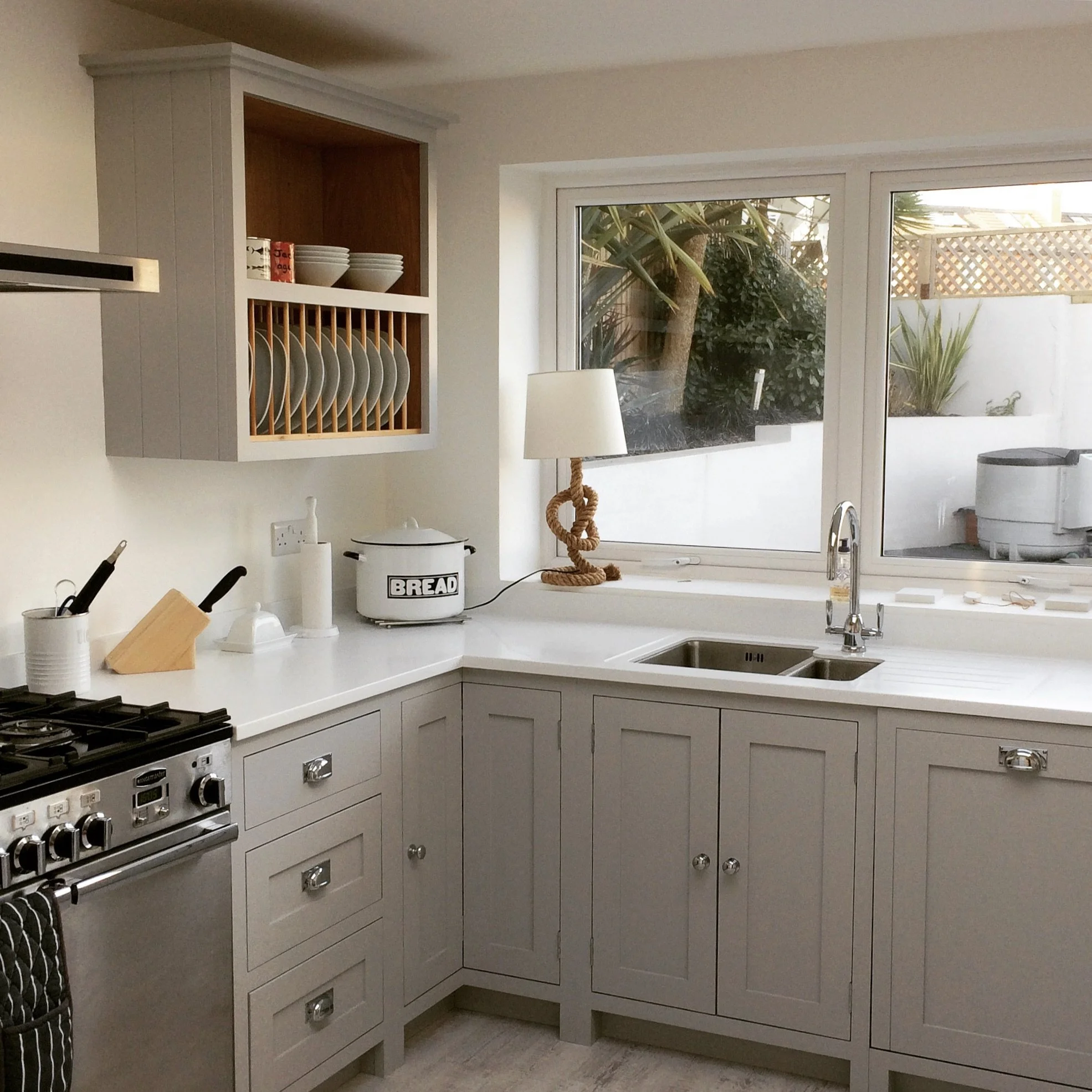 A modern kitchen with light gray cabinets, white countertops, a double sink under a large window, and a stove. Open shelving above holds dishes and bowls. On the counter are a bread box, paper towel holder, and knife holder. There is a decorative lam