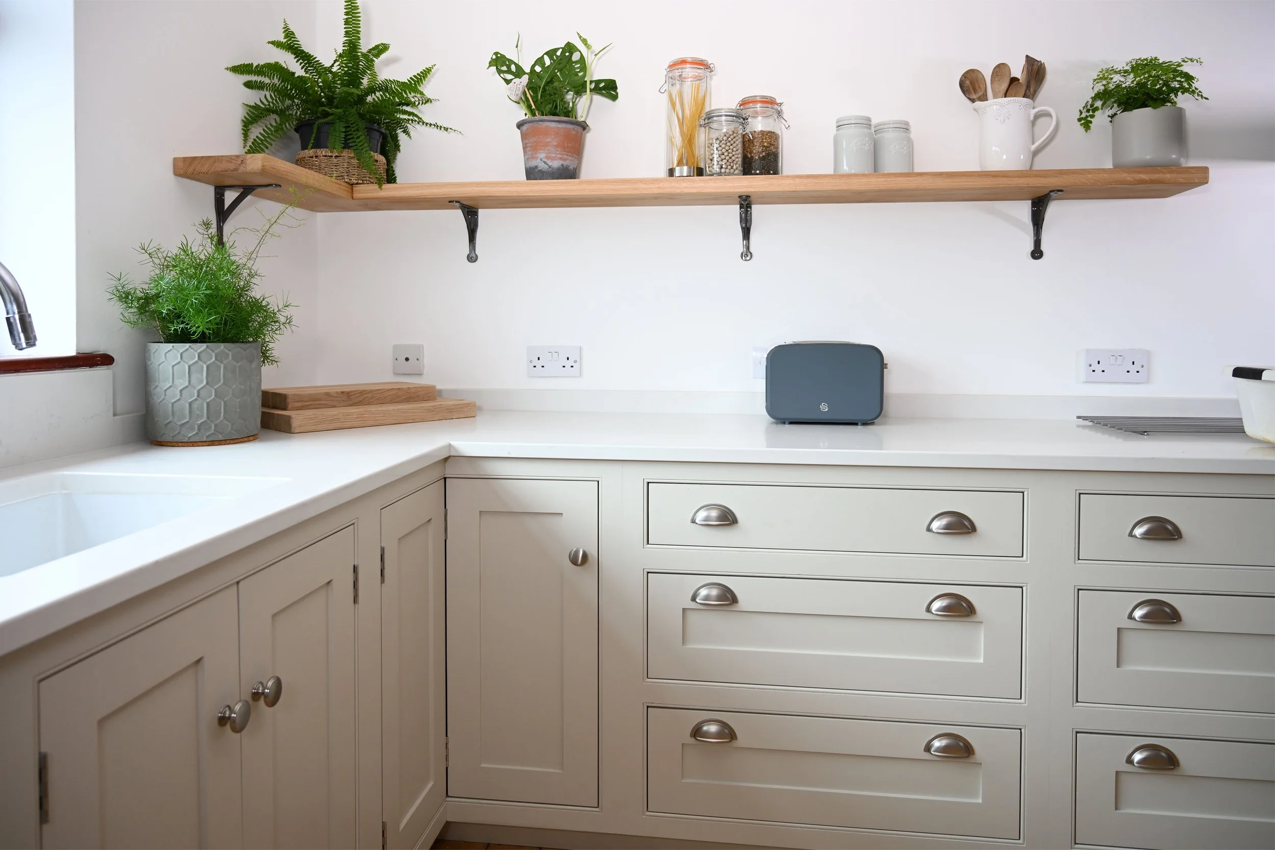 Kitchen with white cabinetry, a white countertop, a potted green plant on the left, kitchen utensils and jars on a wooden shelf, and a small toster on the counter.