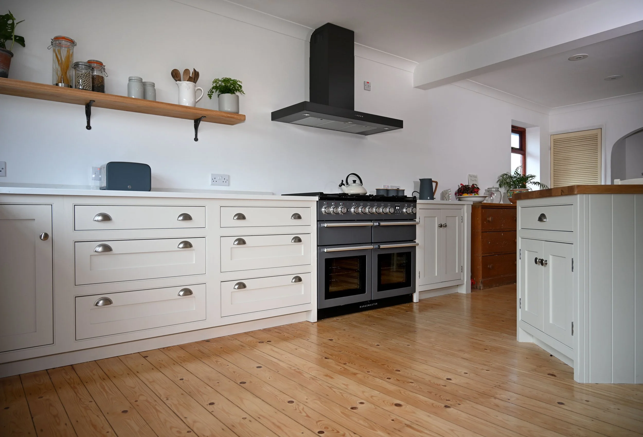Modern kitchen with white cabinets, black stove, wooden countertops, open shelving with jars and plants, hardwood floor, and small window.