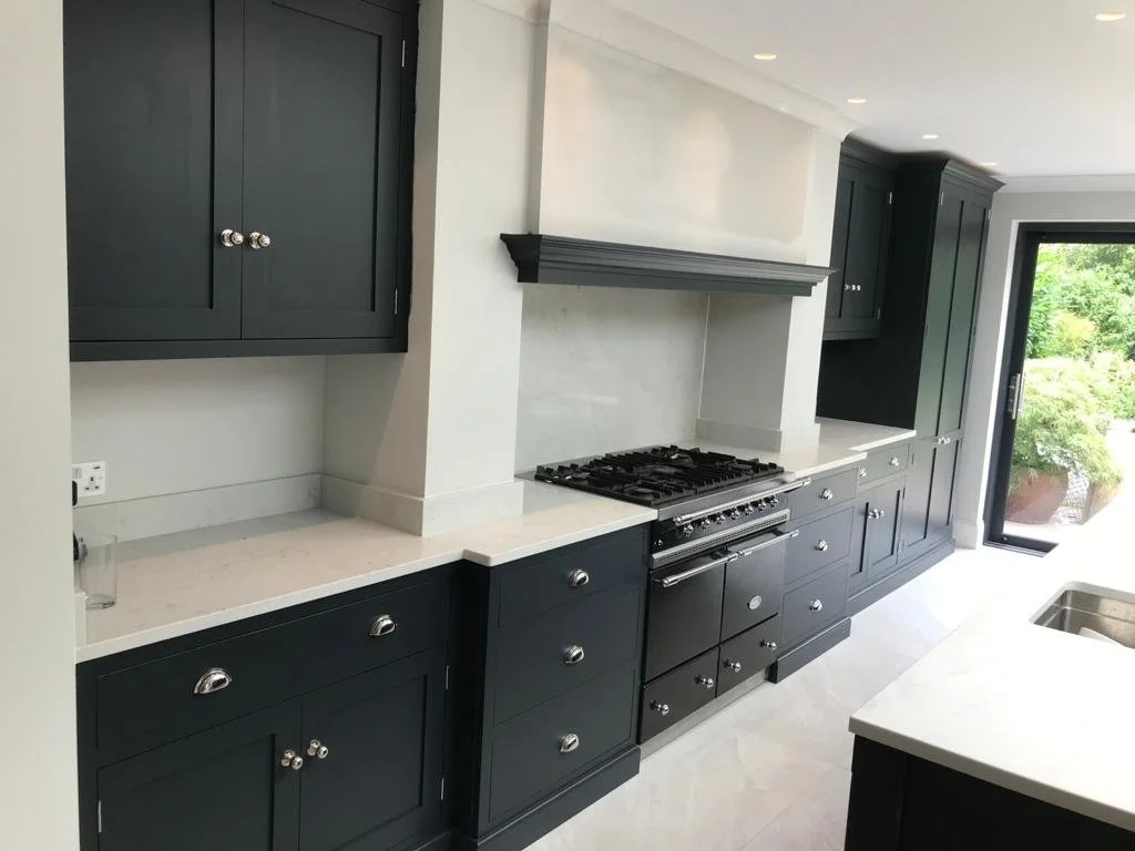 Modern kitchen with dark navy cabinets, white countertops, and a built-in gas stove with a range hood. There is a sliding glass door with greenery outside.