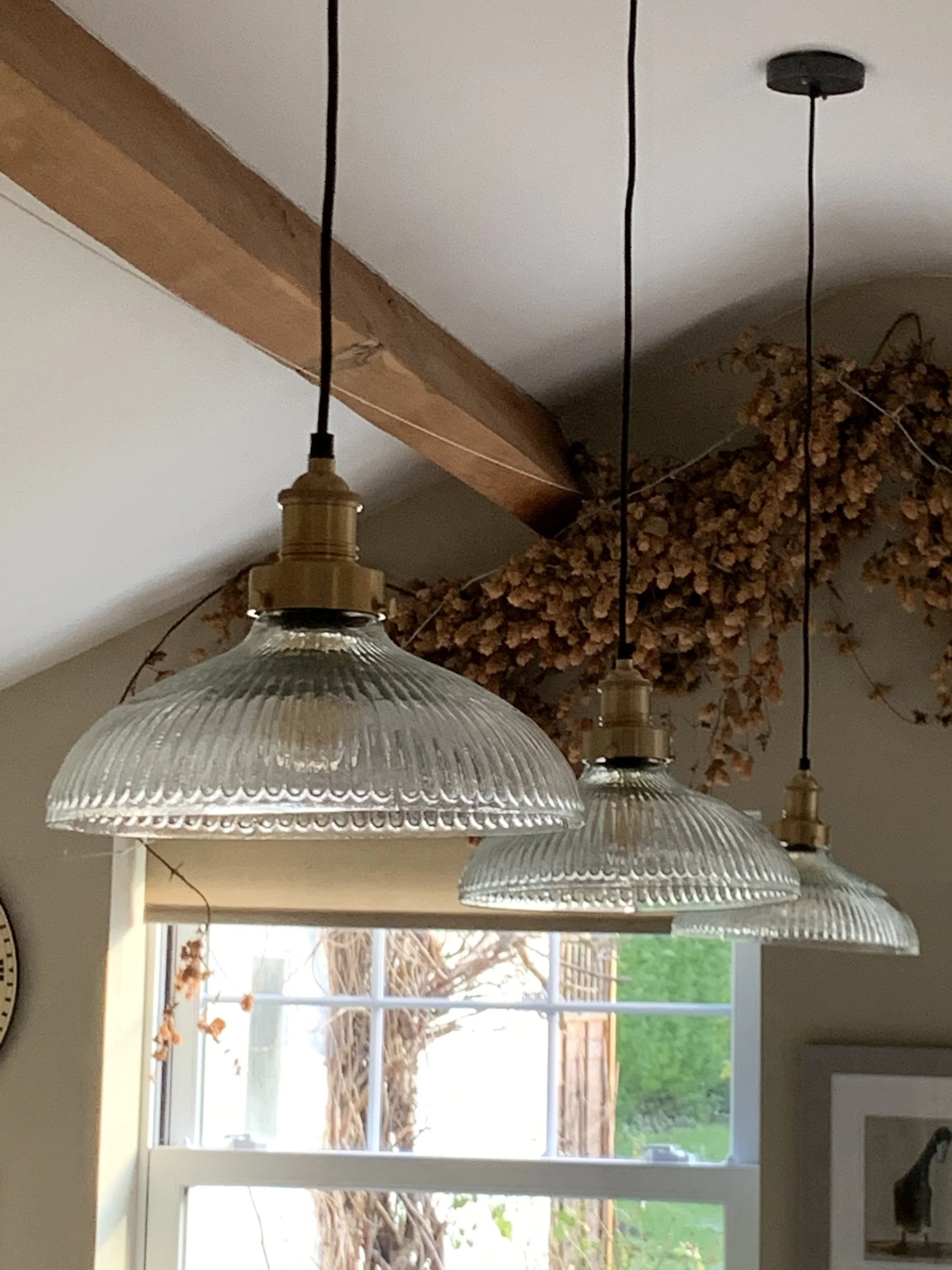 Three glass pendant lights hanging from black cords above a window with dried plant decor and a wooden beam.