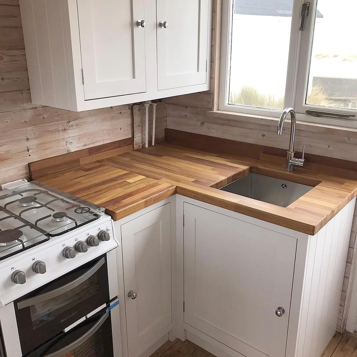 A kitchen corner with wooden countertops, white cabinets, a stainless steel sink, a gas stove, and a window above the sink.