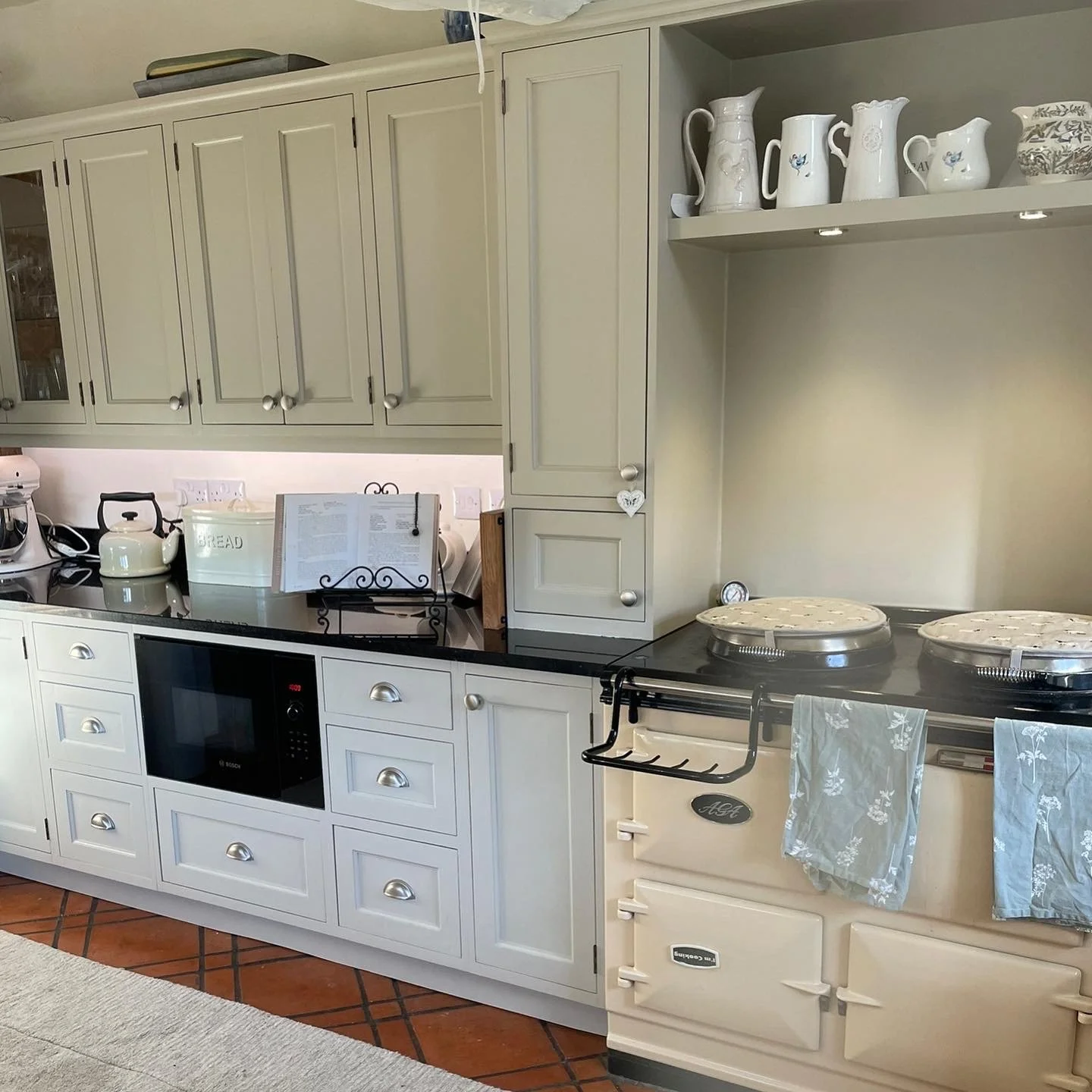 Kitchen with cream-colored cabinets, a black countertop, kitchen appliances including a microwave, and decorative ceramic pitchers on a shelf. An Aga stove with towels hanging on its handle is also visible.