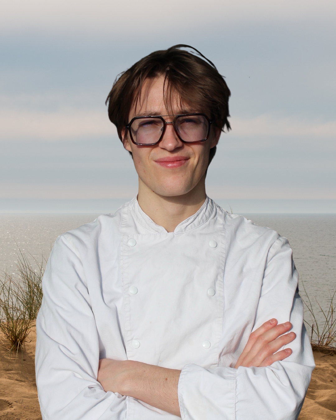 Person dressed as a chef in white coat with crossed arms standing on a sandy beach near the ocean, with dunes and grass behind.