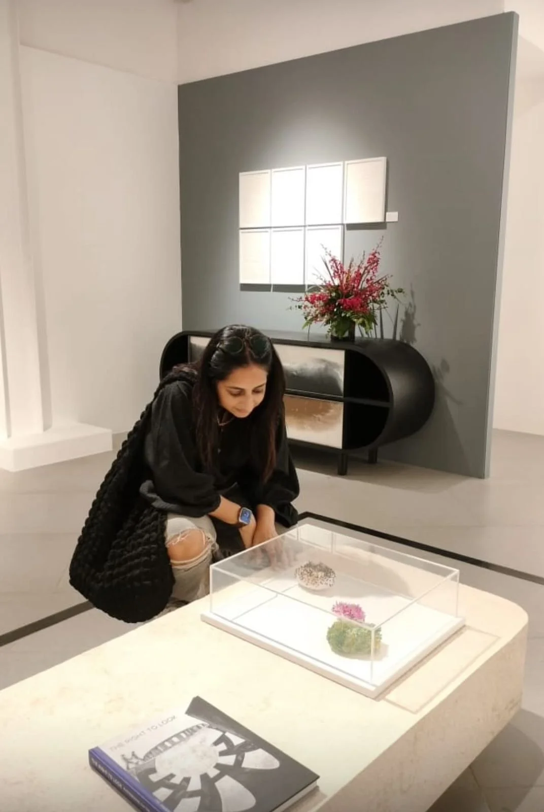 A woman with dark hair wearing a black sweater, ripped jeans, and a smartwatch, crouches down to look at jewelry in a display case inside a modern art gallery or museum. Behind her, there is a black sideboard with a flower arrangement on top, a back wall with a small window reflection, and a gray wall panel.