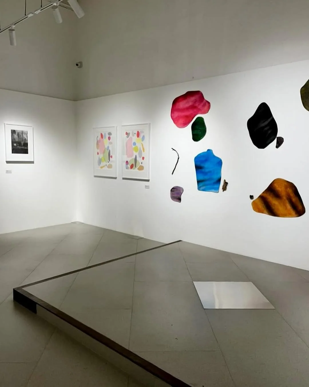 An art gallery with white walls and tiled floors displaying framed paintings and abstract sculptures of colorful, irregularly shaped pieces on the wall.