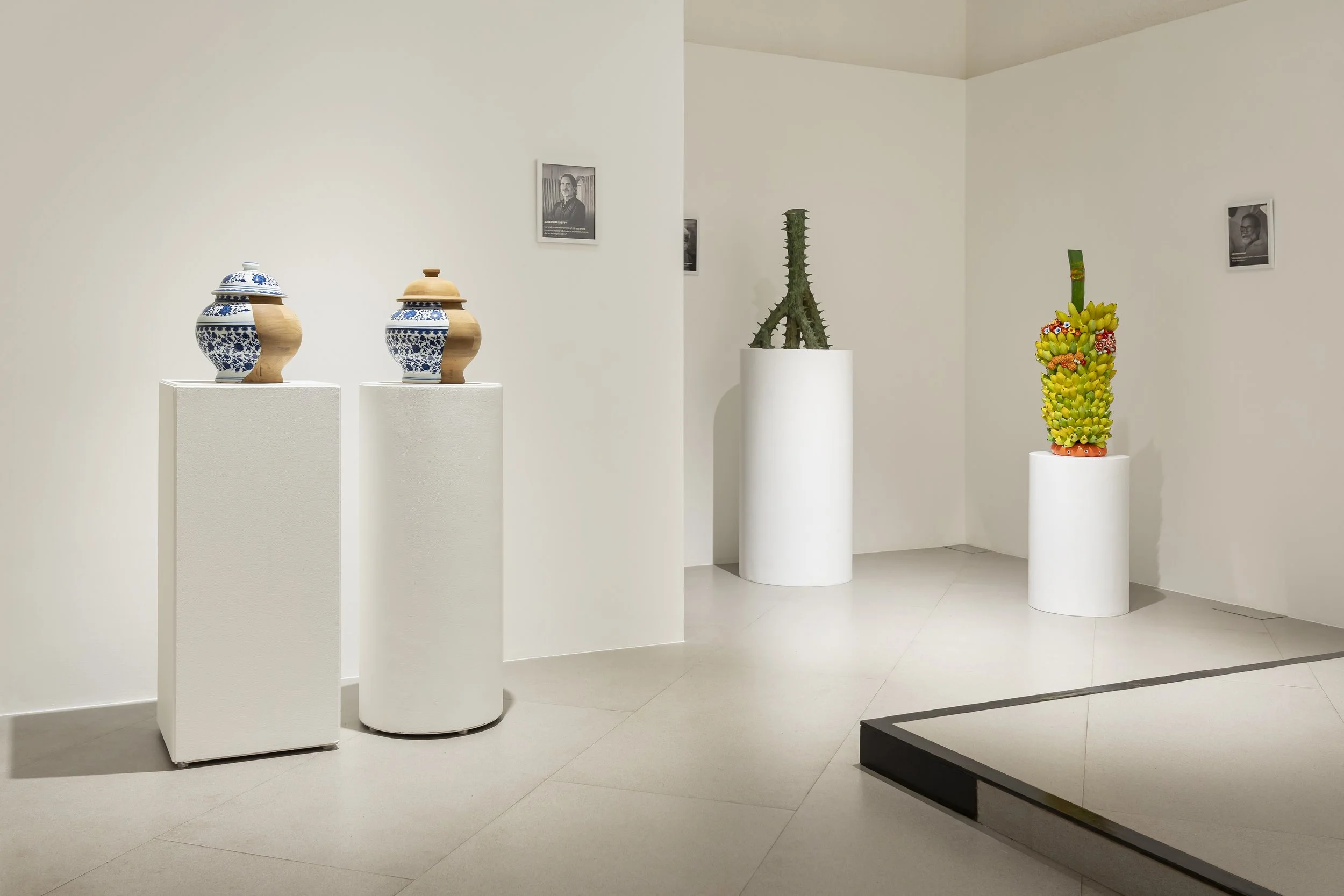 Art gallery exhibit featuring two blue and white ceramic jars with wooden lids on white pedestals to the left, a tall cactus plant on a white pedestal in the middle, and a colorful ceramic sculpture resembling a fruit bouquet on a white pedestal to the right, with framed black-and-white photographs on white walls.