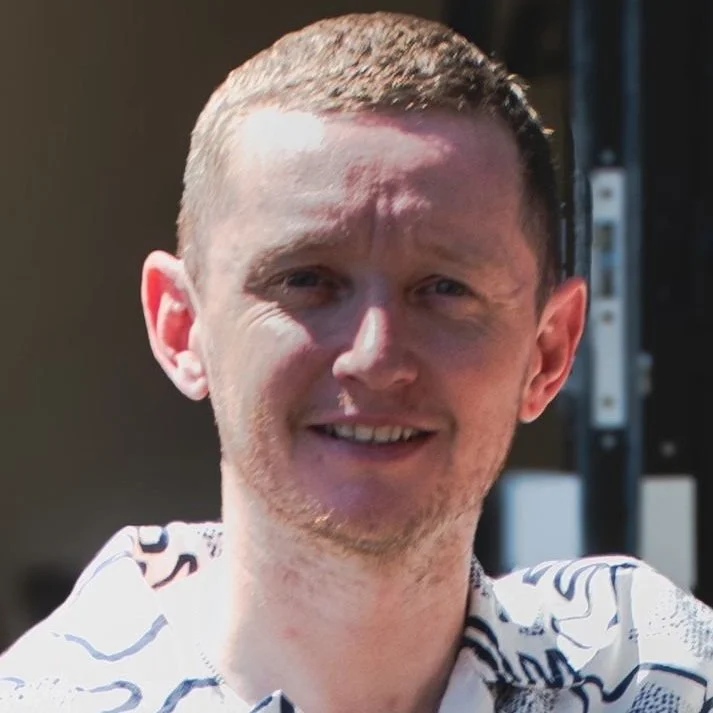 A young man with short light brown hair and a light beard, smiling outdoors on a sunny day.