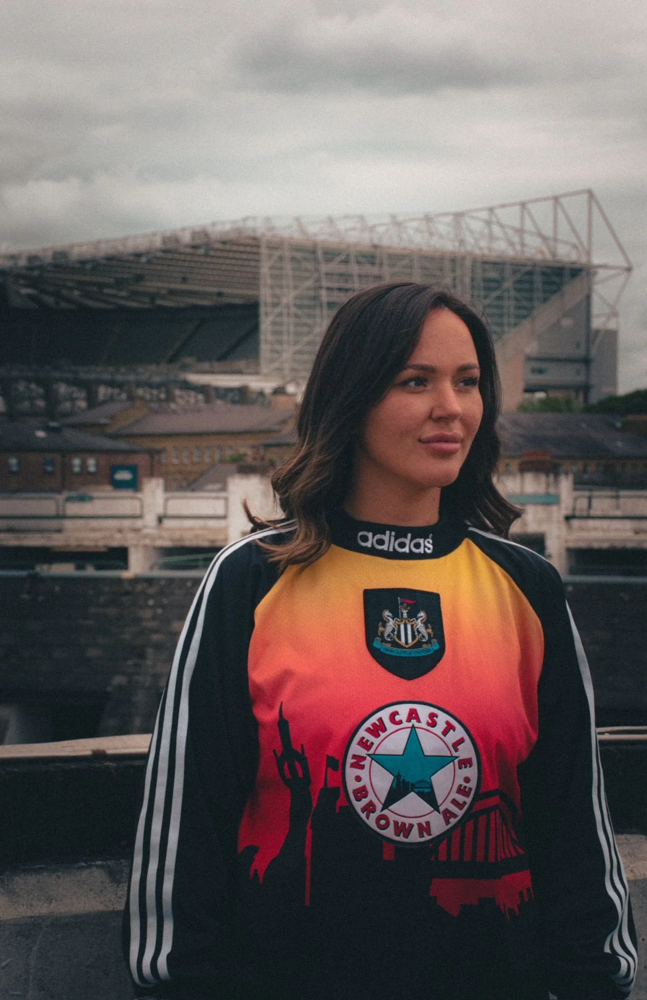 A woman in a colourful Newcastle United football jersey shirt outdoors with a stadium in the background on a cloudy day.