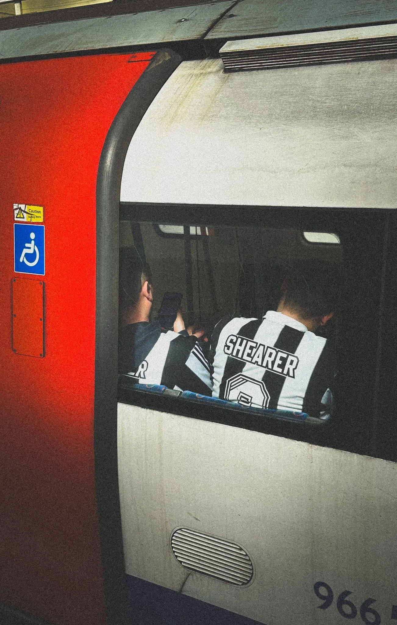 Two people sitting on a London Underground carriage, one looking at a phone, wearing black and white Newcastle United shirts, one with the name 'Shearer' on the back.