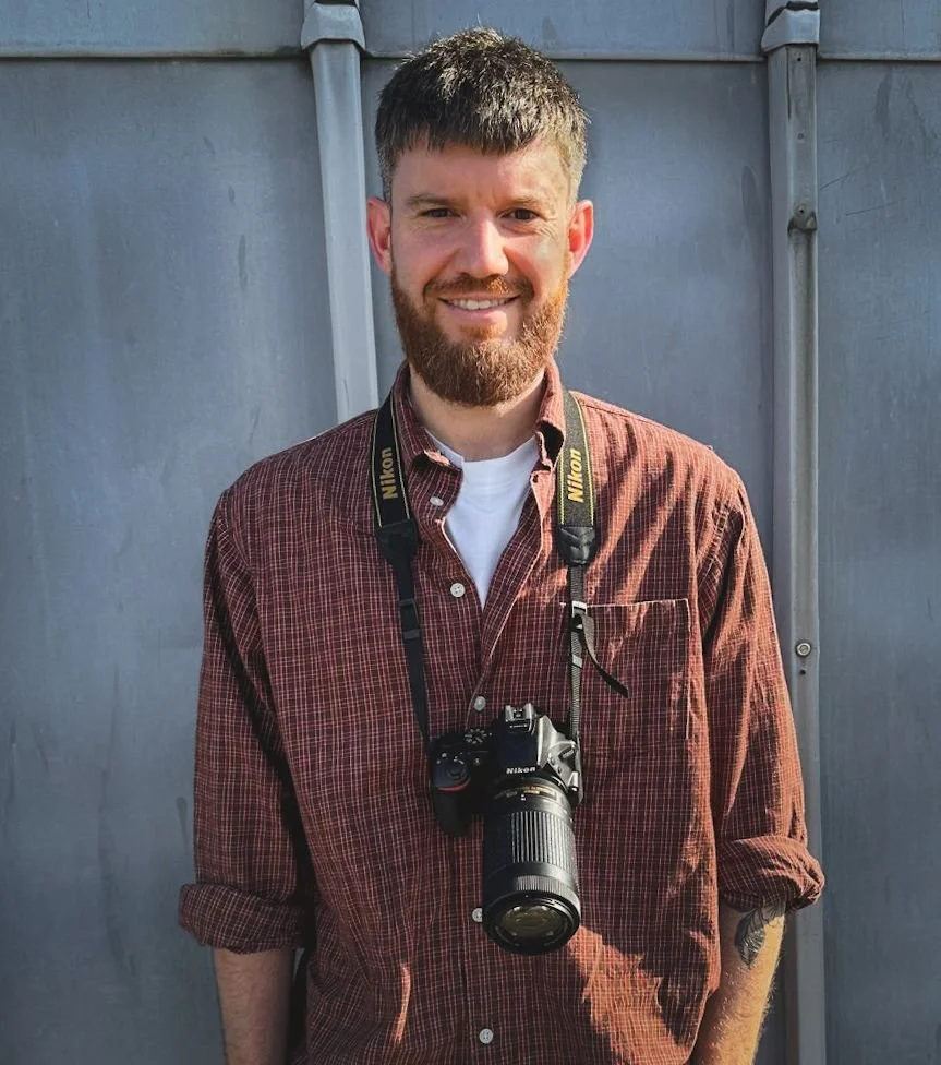 A smiling man with a beard and short hair wearing a red checkered shirt with sleeves rolled up, standing outdoors in front of a gray wall, with a Nikon camera hanging around his neck.