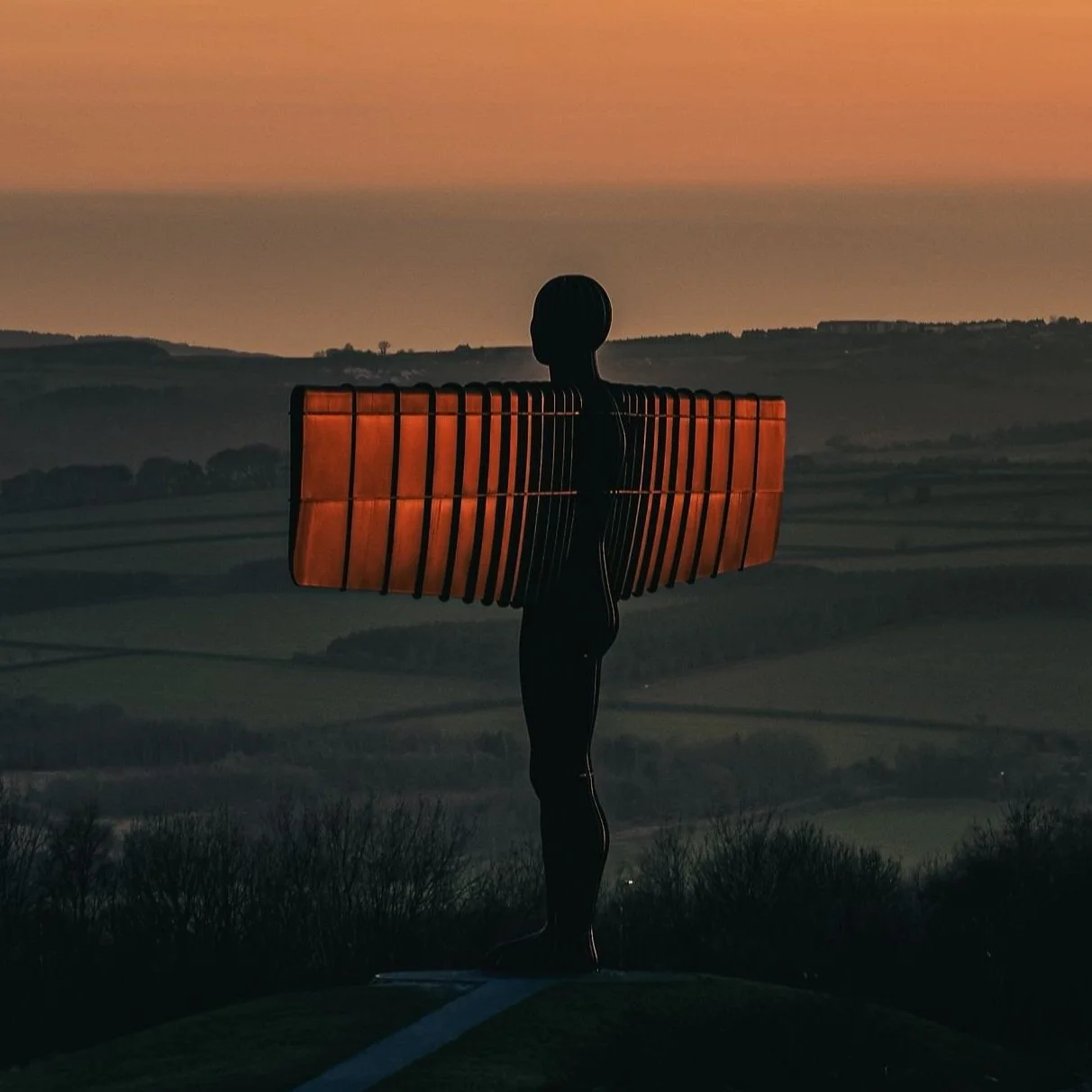 Sculpture of a human figure with wings made from metal, silhouetted against a sunset over rolling hills.