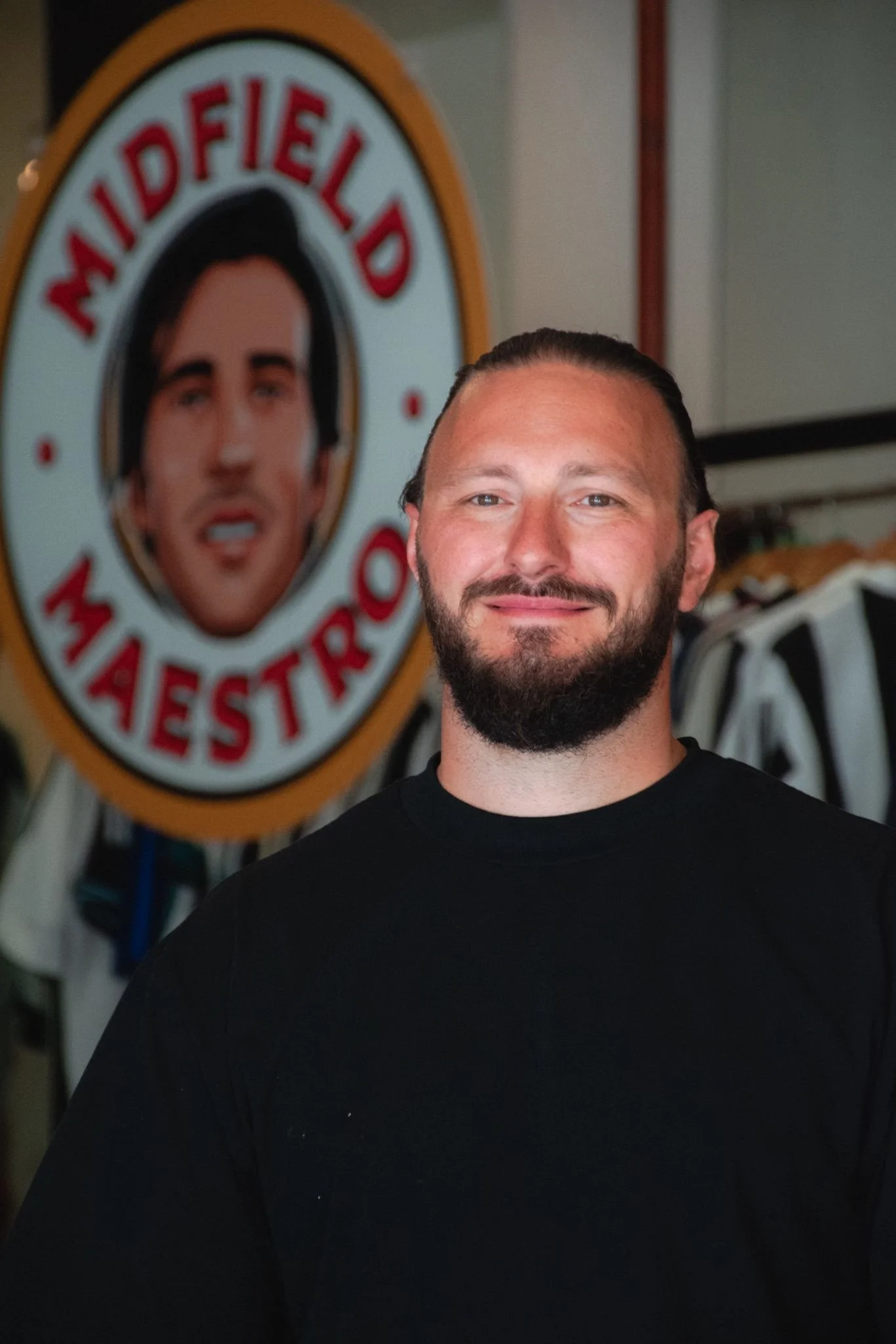 A man with a beard standing in front of a sign that says 'Midfield Maestro' with a portrait of a man with dark hair in the background.