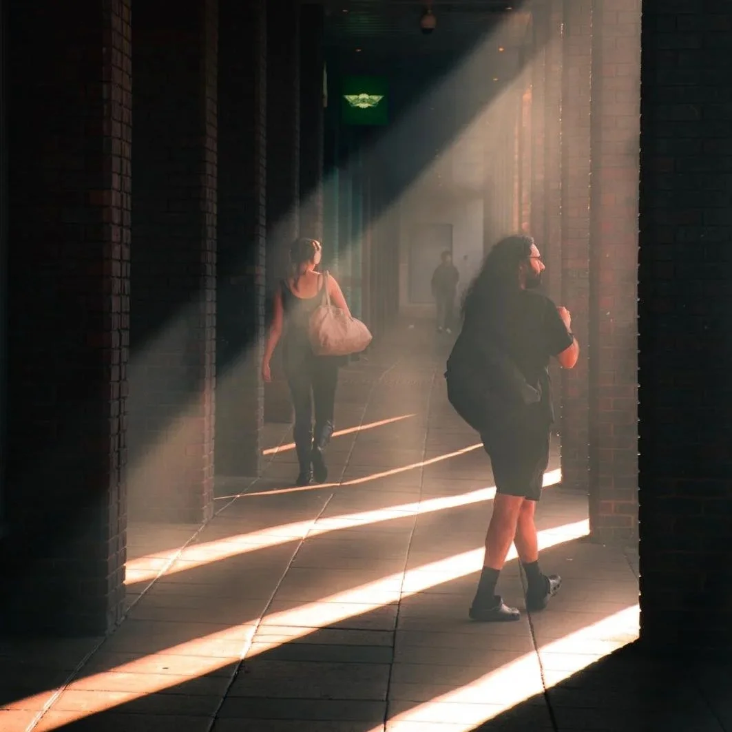 Sunlight streams through a walkway with brick walls. Three people walk through: a woman with a large shoulder bag walking away, a man with a backpack standing near the wall, and another person in the background.