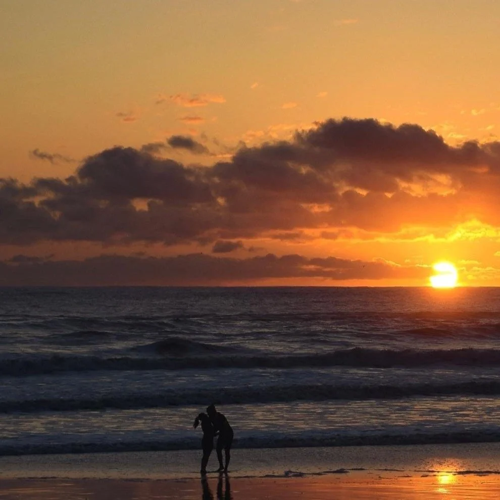 Silhouettes of two people on the beach at sunset, with the sun partly below the horizon and clouds in the sky.