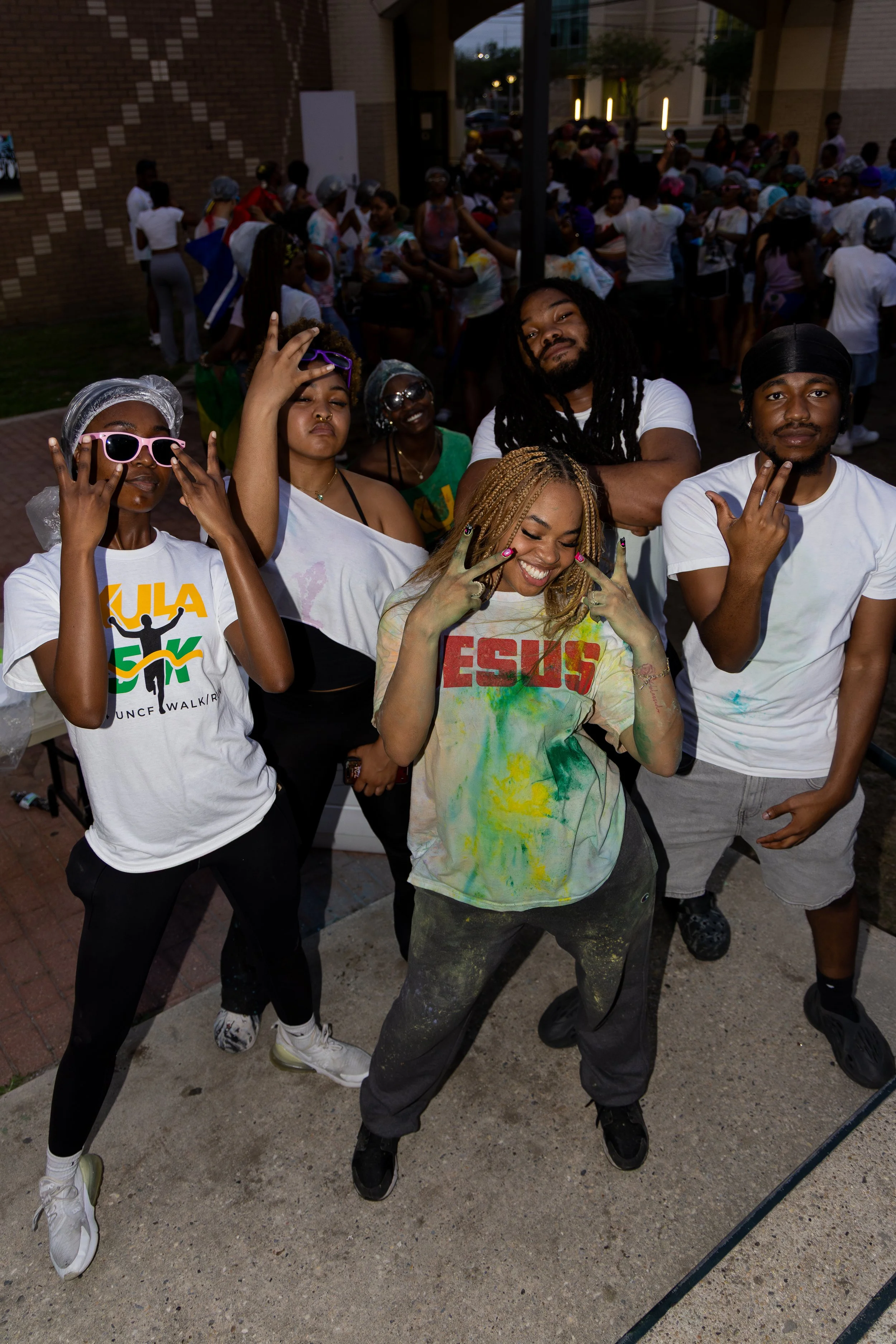 Group of young people at a color run event, posing for a photo with colorful powder on their clothes and hands, with a crowd in the background.