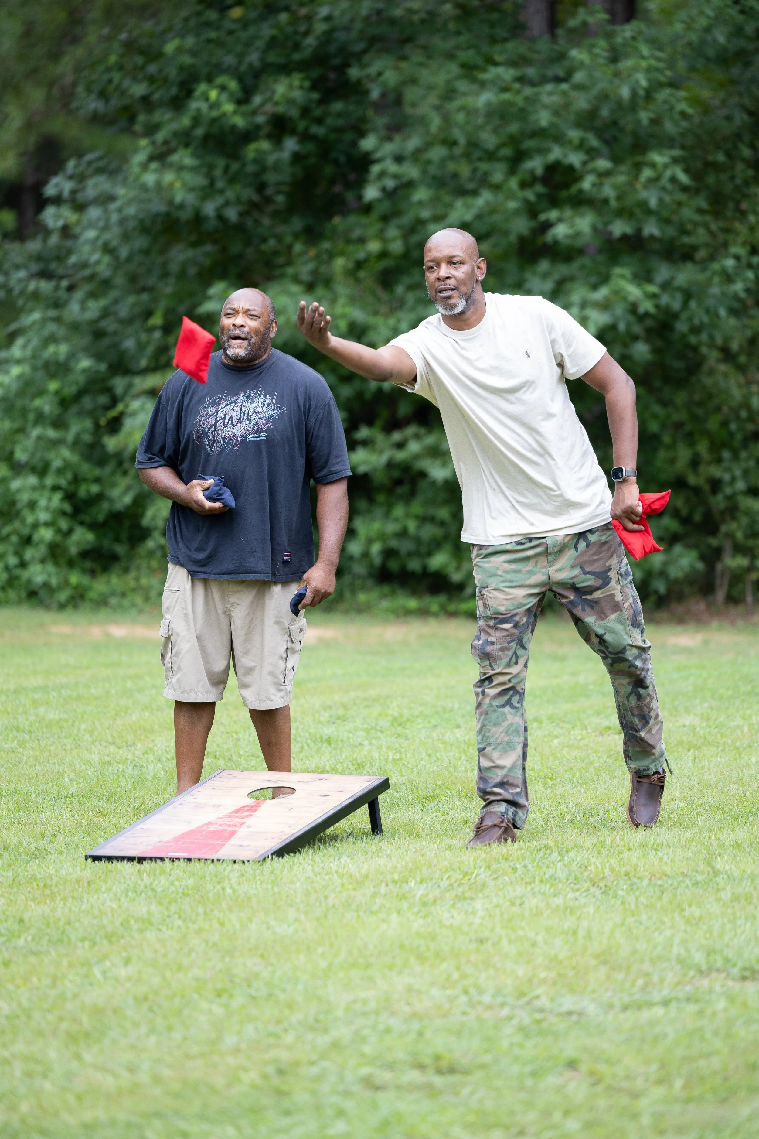 Two men playing cornhole outdoors on a grassy field, one throwing a bean bag while the other observes, with trees in the background.