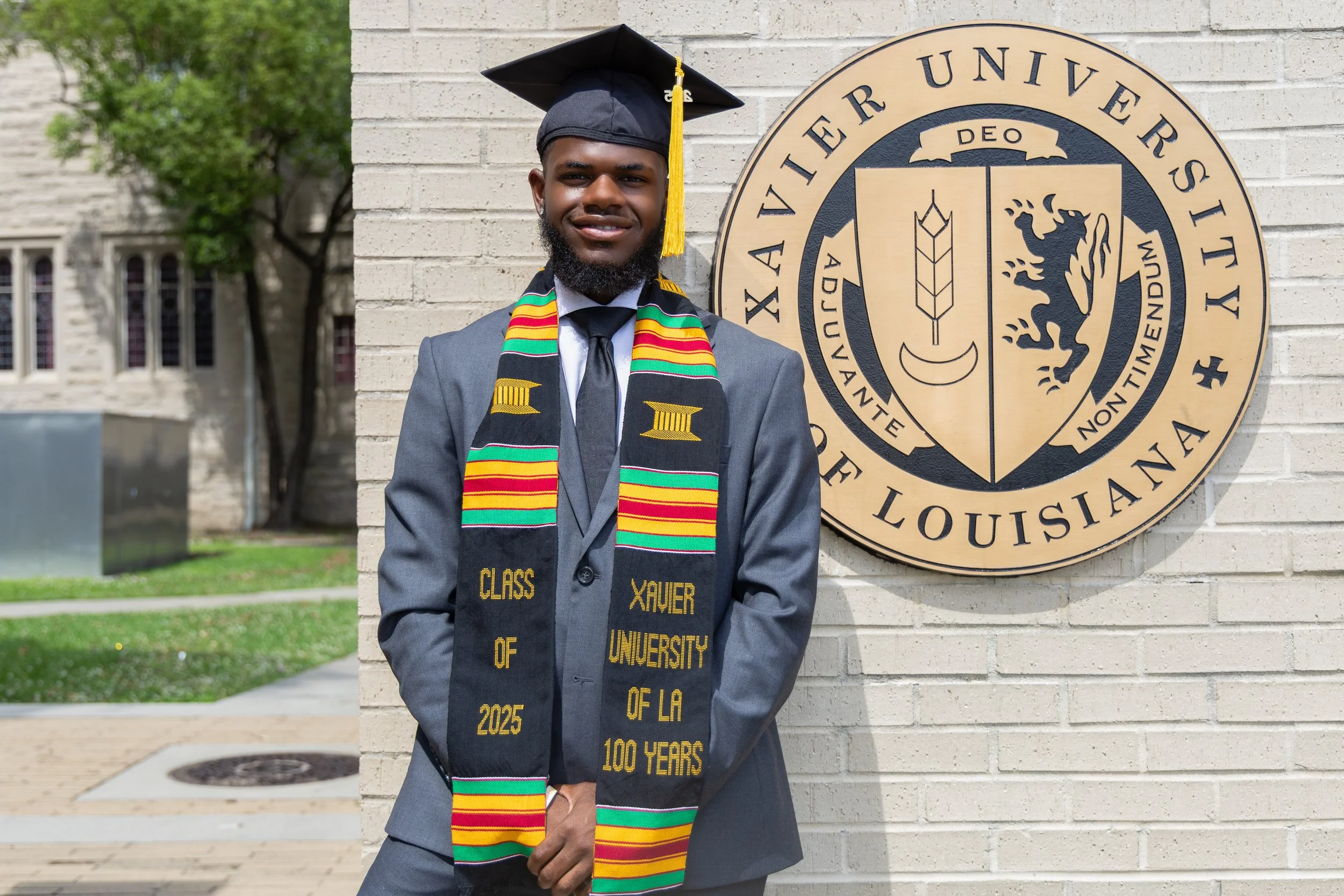 A young man in a graduation cap and gown stands outside near a brick wall with the Xavier University of Louisiana seal. He is smiling, wearing a colorful stole with the text "Class of 2025" and "Xavier University of LA 100 Years."