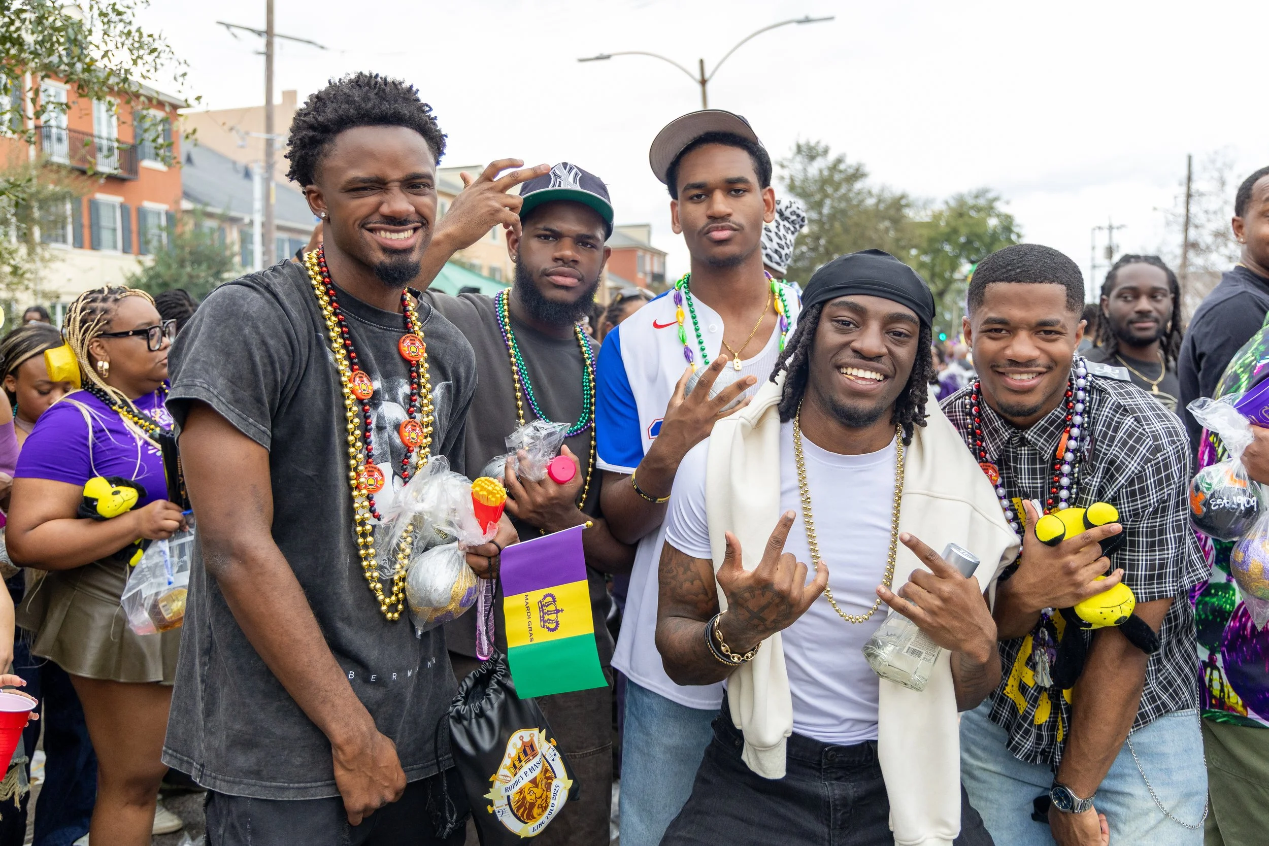 Group of young men smiling and making hand gestures at an outdoor event, wearing necklaces and casual clothing.