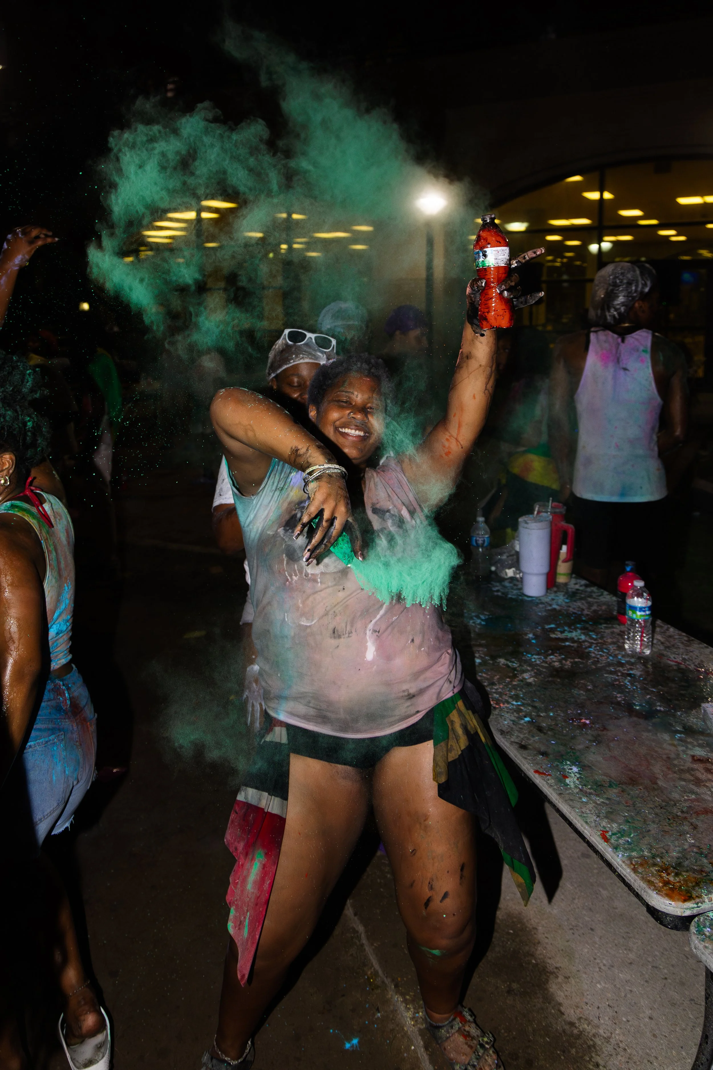 A woman celebrating at a color festival throws colored powder into the air while smiling and holding a beverage, surrounded by other participants covered in colorful powders at night.