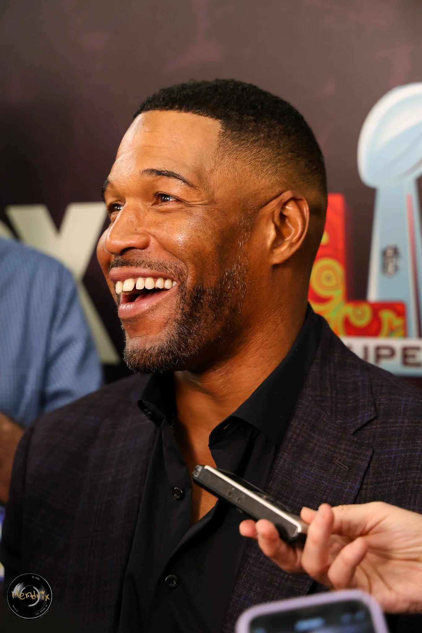 Man with beard smiling and speaking into microphones, wearing a dark suit and black shirt, at a media event.
