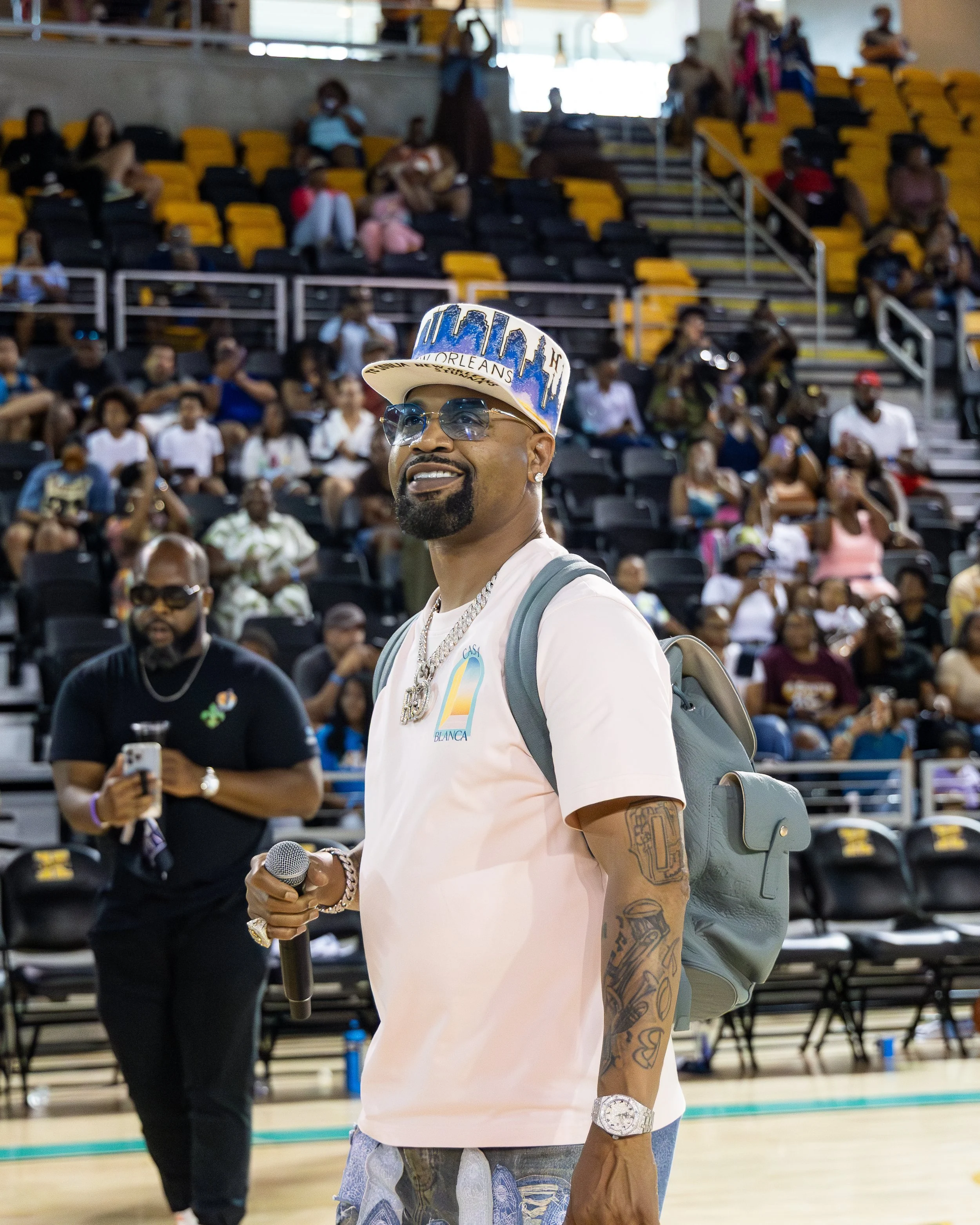Rapper Juvenile wearing sunglasses, a hat with 'New Orleans' written on it, and a pink t-shirt , holding a microphone, standing in a sports arena with a crowd in the background.